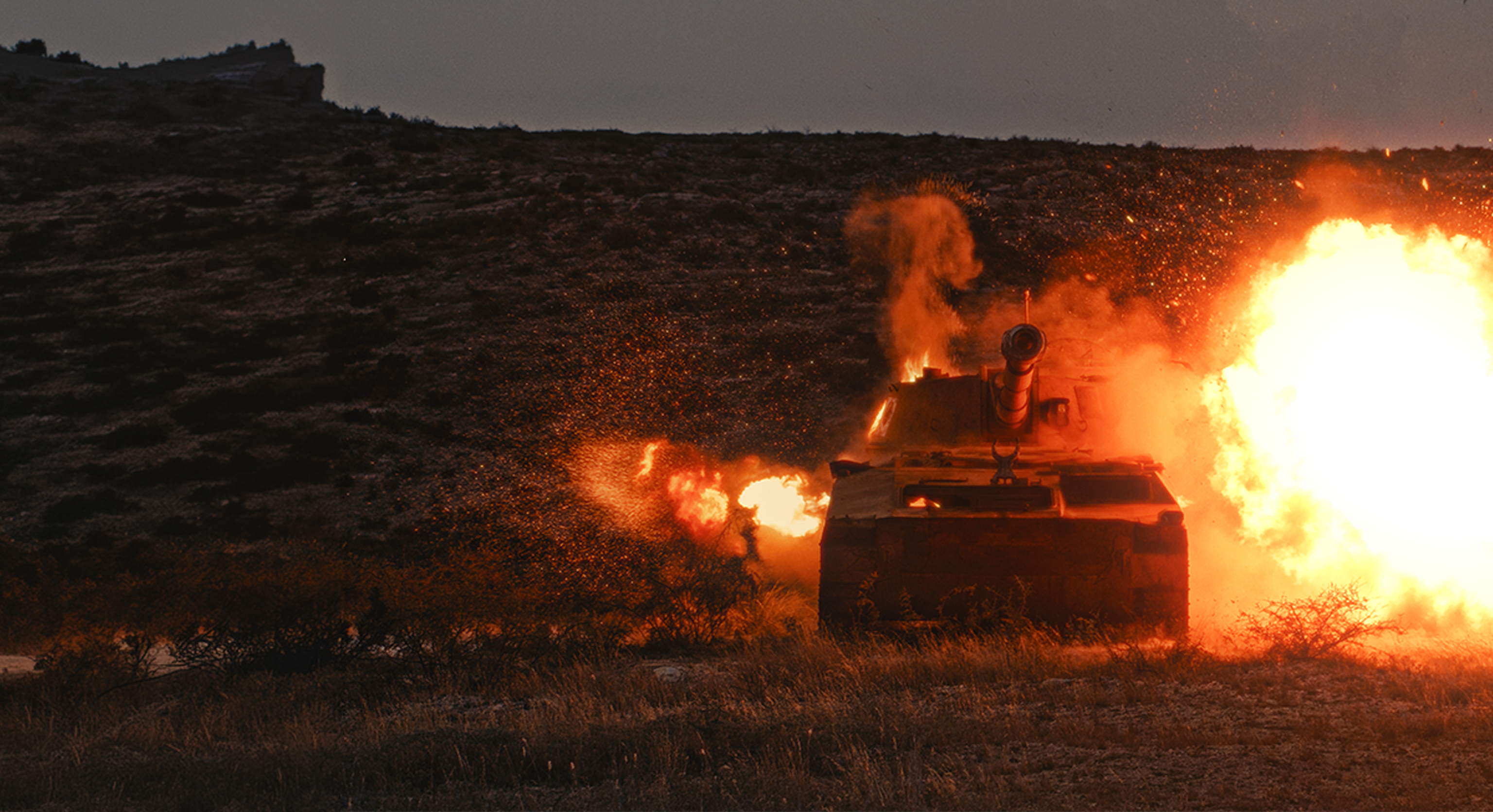 Front view of a tank firing a large explosive round in a dry, rugged landscape during dusk or dawn, with bright flames and smoke bursting from the barrel.
