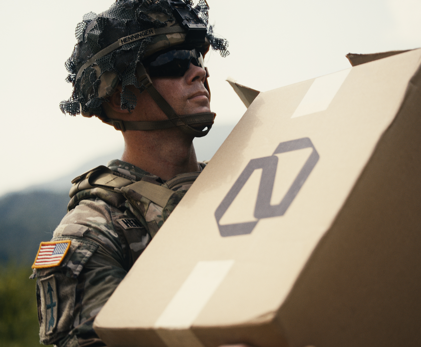 Military personnel in camouflage uniform and helmet carrying a large cardboard box with a printed logo, outdoors with blurred natural background.