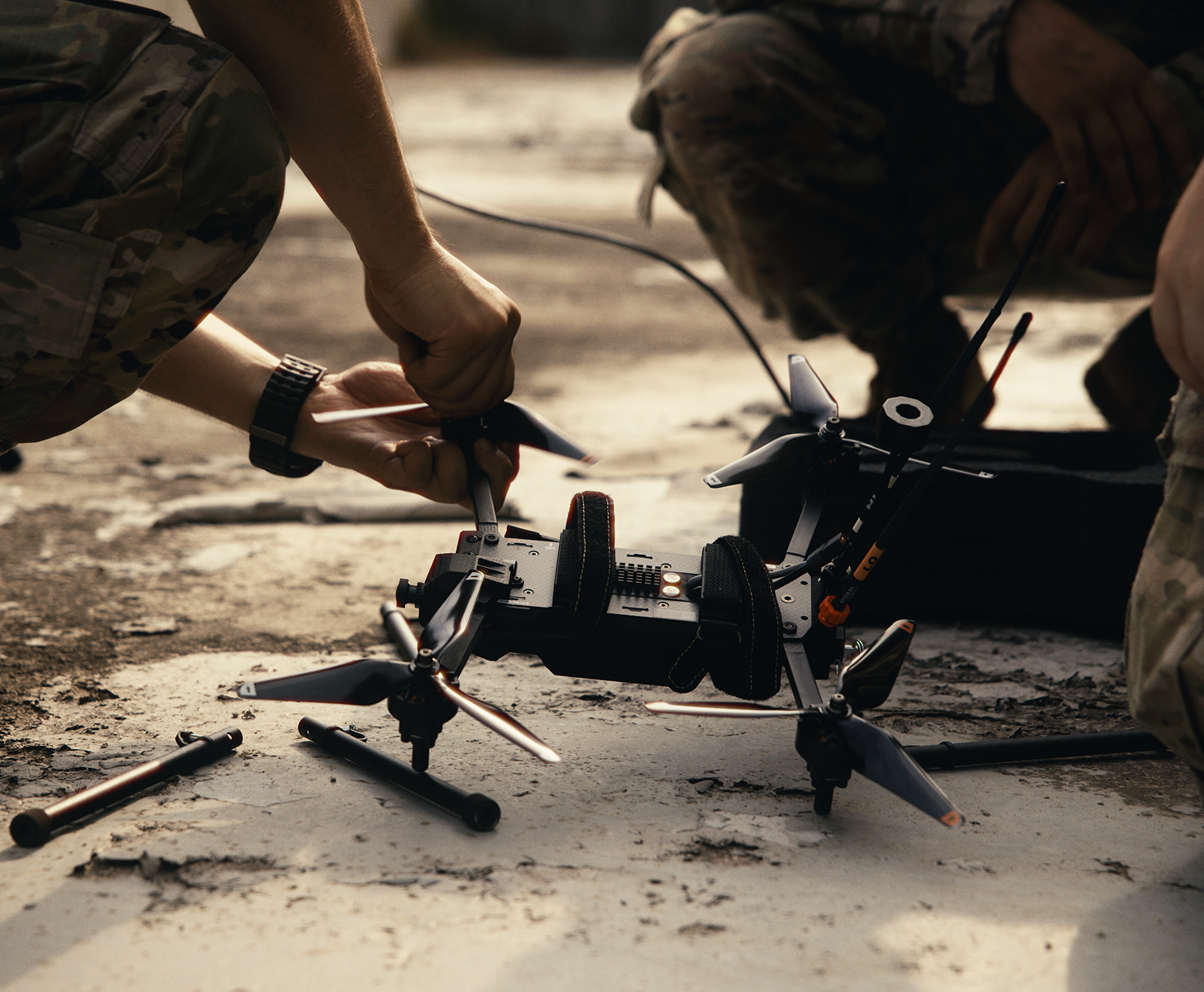 Two individuals in camouflage uniforms kneeling while assembling or repairing a black quadcopter drone on a rough surface.