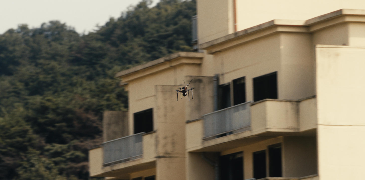 Small black drone flying near a beige building with balconies, with a backdrop of dense green trees.