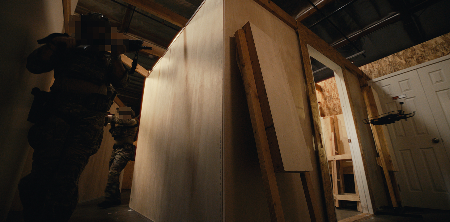 Two armed individuals in tactical gear move cautiously through a dimly lit indoor wooden structure, while a black drone hovers near an open doorway with white doors and wooden frames.