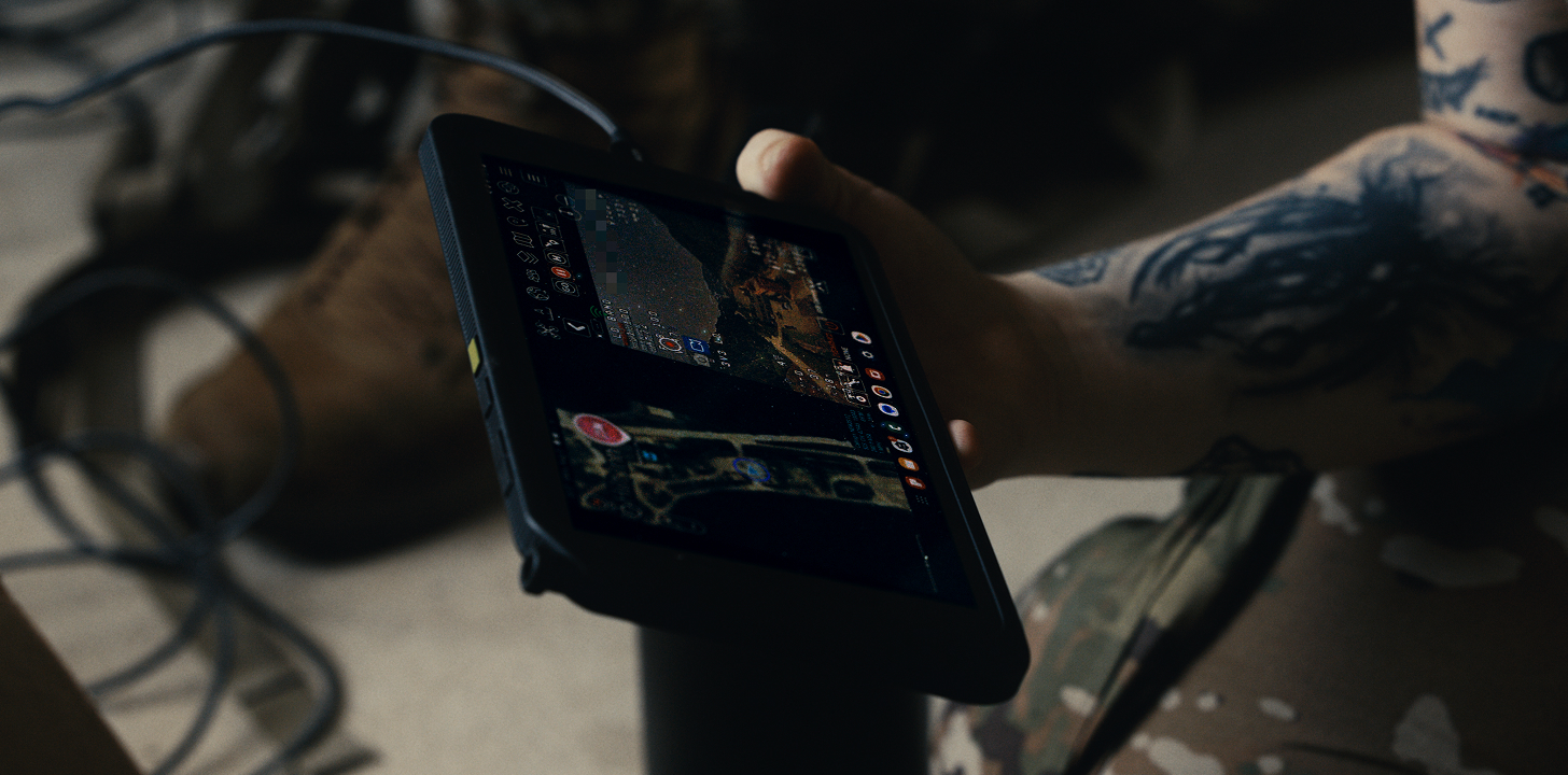 Person with tattooed arm holding a rugged tablet device displaying a tactical map and data, wearing camouflage pants and brown boots, with cables on the ground.