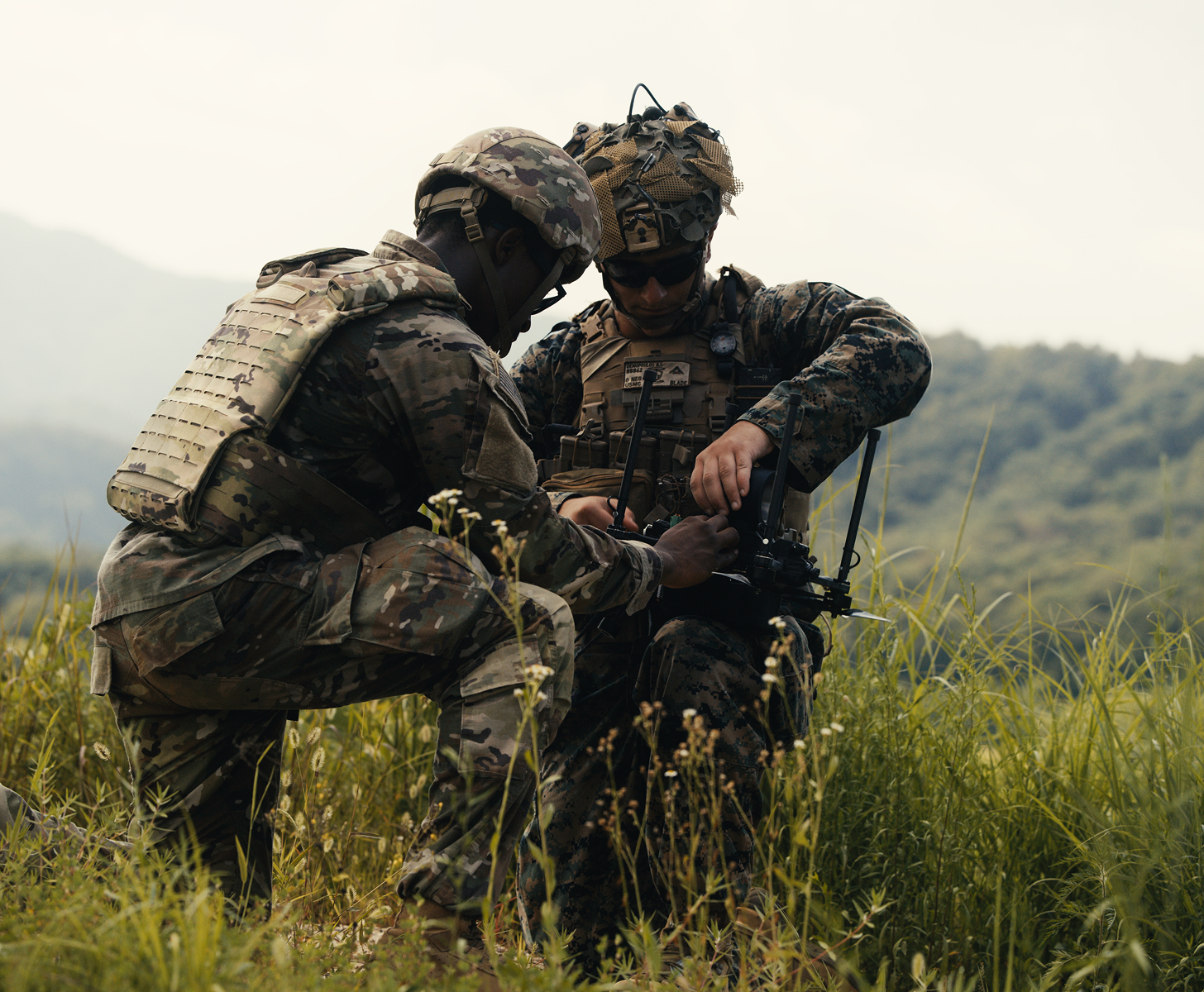 Two soldiers in camouflage uniforms and tactical gear are kneeling in tall grass on a hillside, working together to operate a device with antennas.