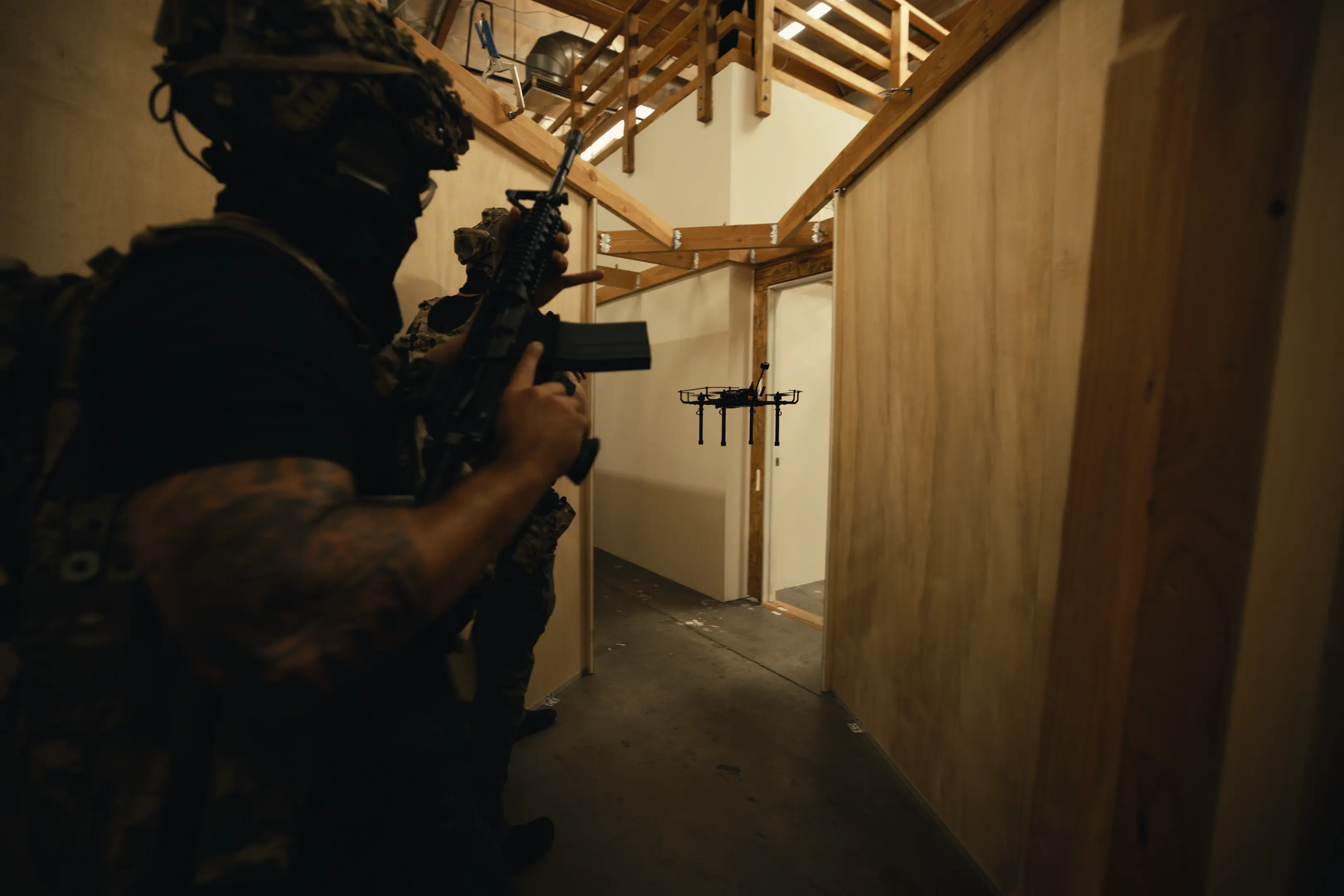 Two soldiers in tactical gear holding rifles inside a building hallway with wooden panel walls, facing a black drone hovering ahead.