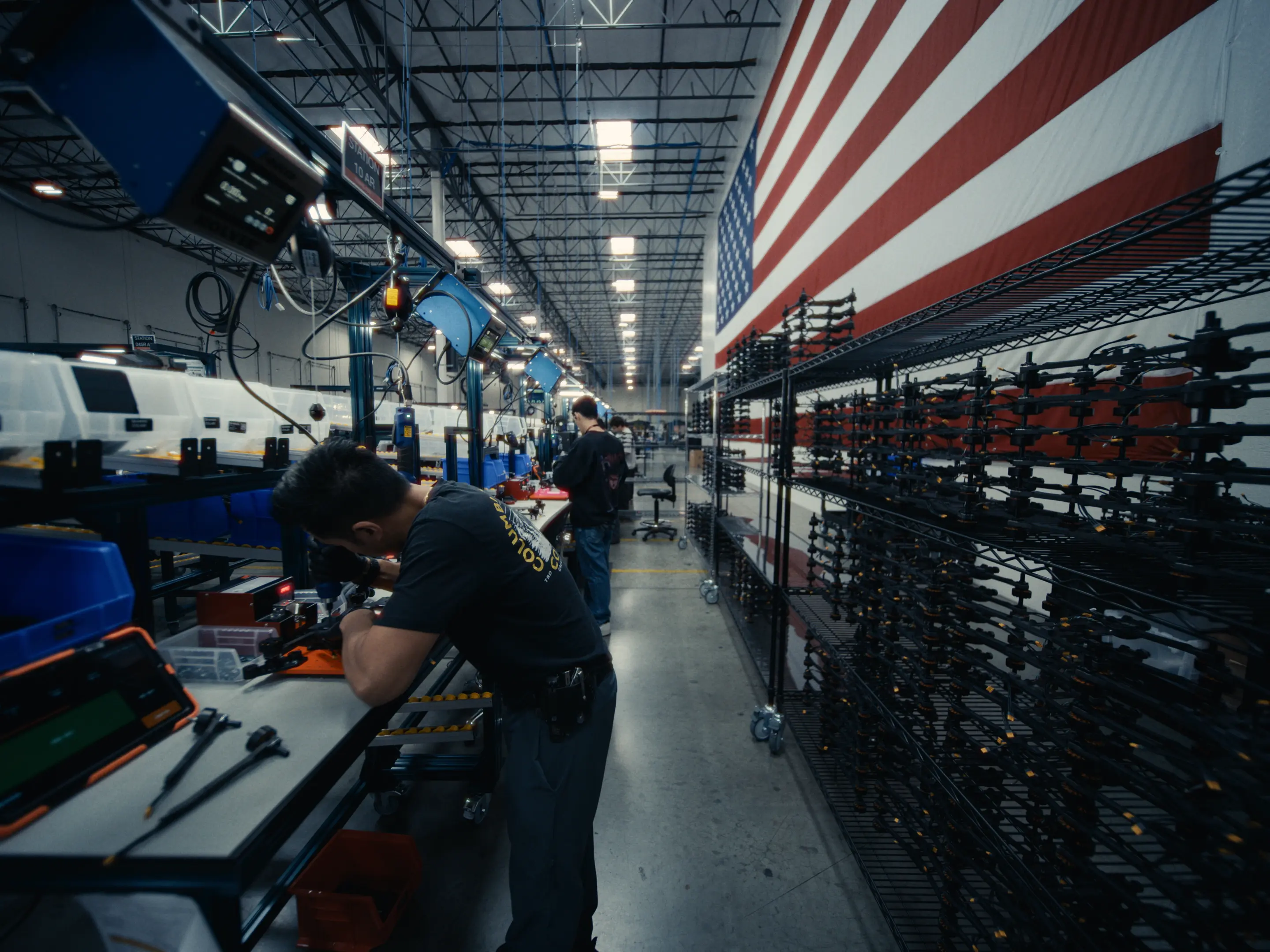 Workers assembling equipment on a production line in a large factory with high ceilings, several workstations, and an American flag on the wall.