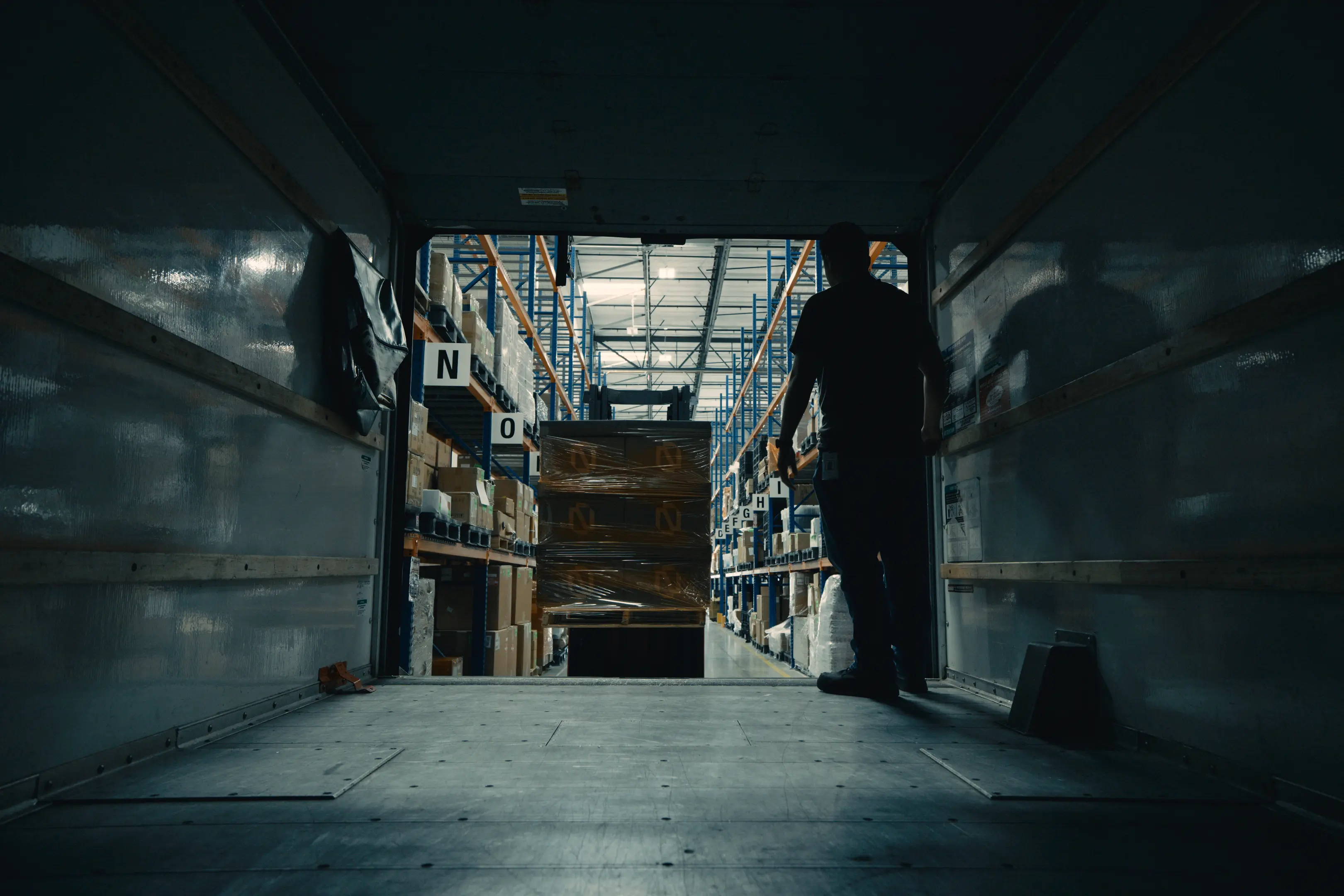 Silhouette of a worker inside a truck trailer facing a warehouse aisle with tall shelves stocked with boxes and a forklift carrying wrapped pallets.