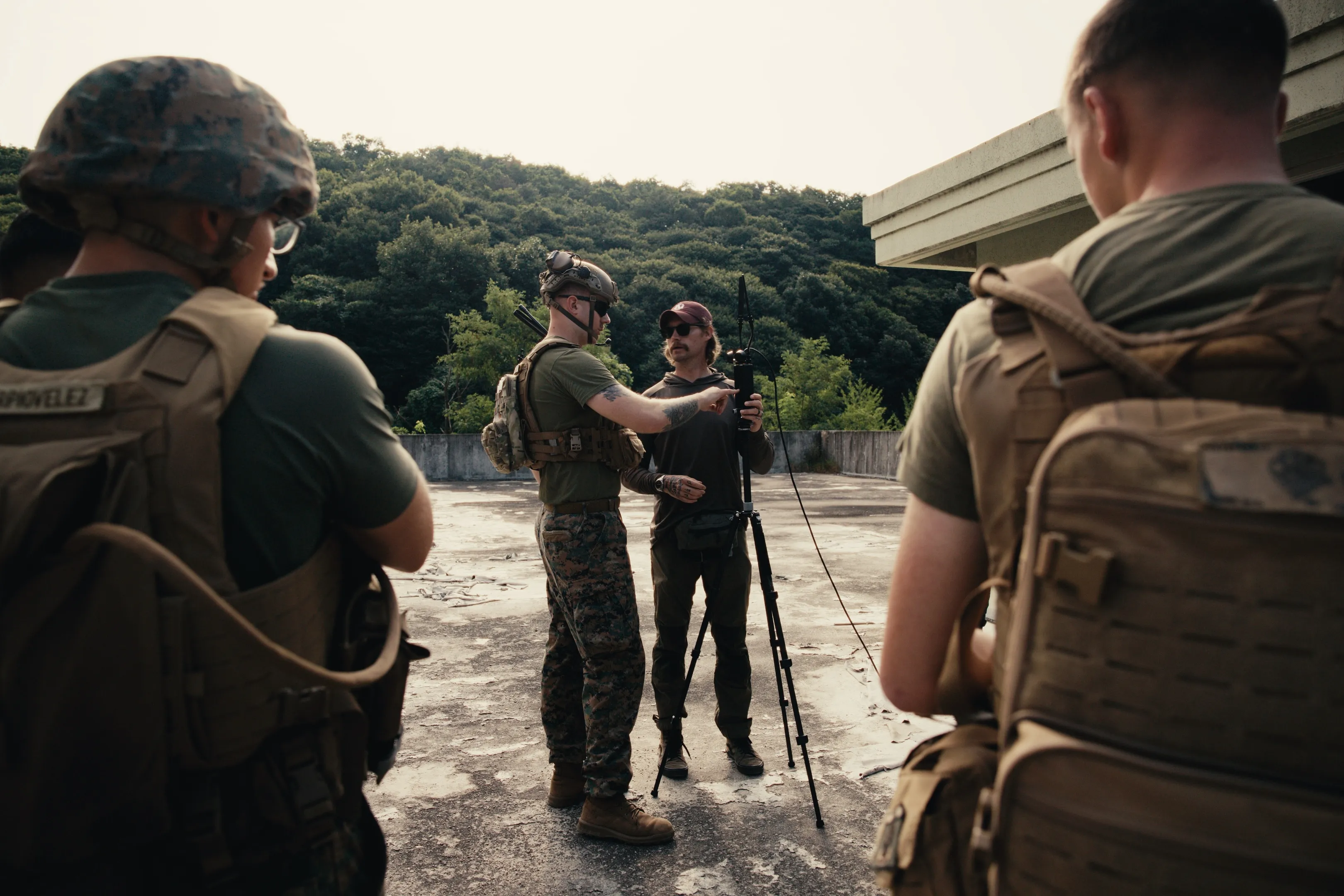 Four people outdoors on a concrete surface near a building, three wearing military gear and one in casual outdoor clothing adjusting a piece of equipment mounted on a tripod, with wooded hills in the background.