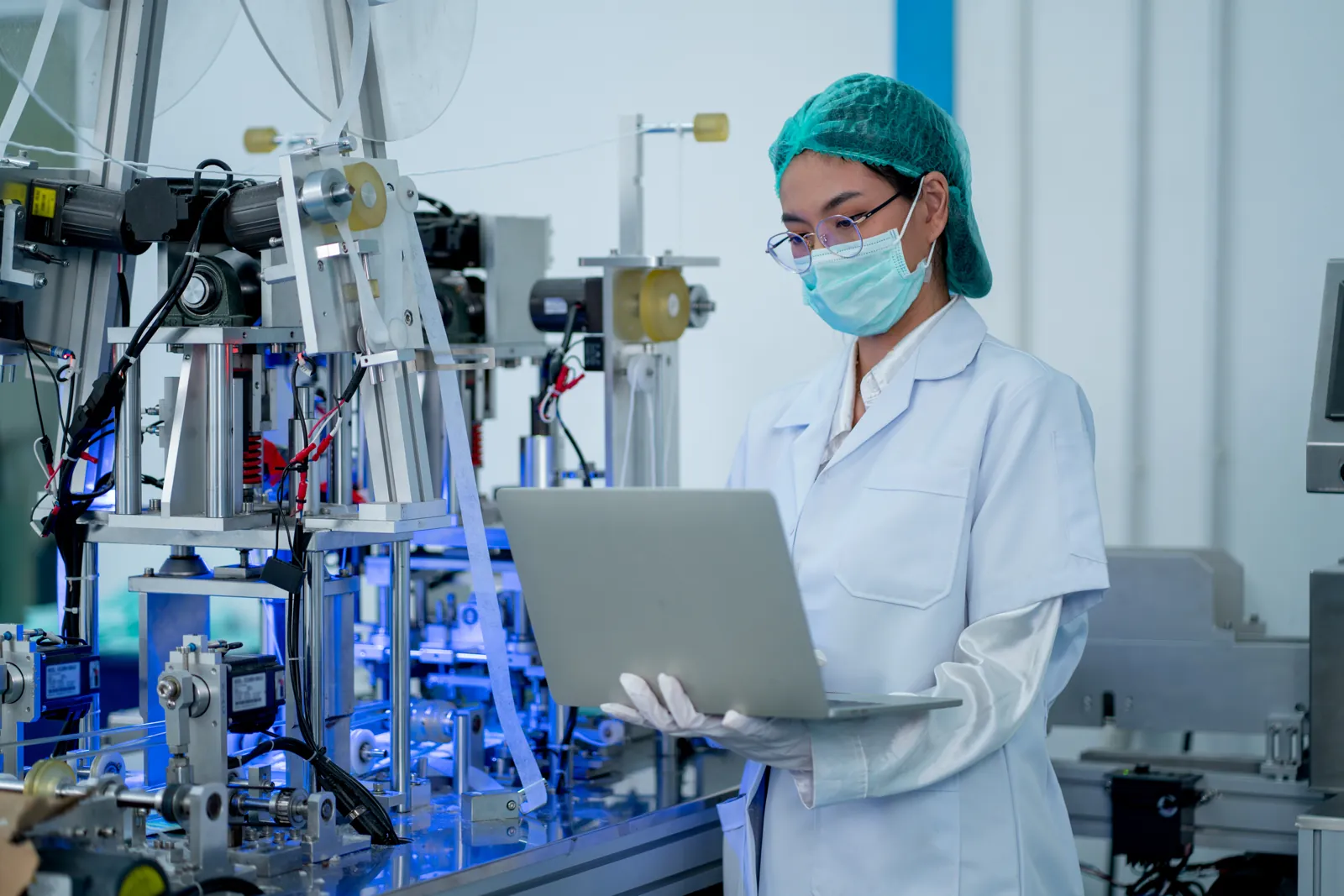 Engineer in a hairnet and mask working on a laptop beside a medical device production line