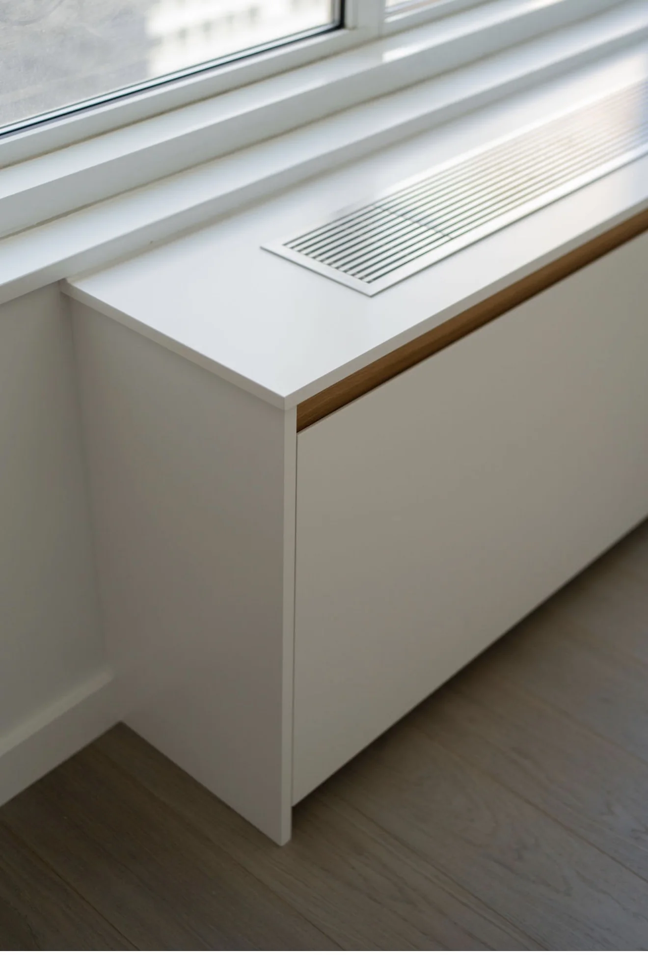 Full-length white radiator cover with a recessed metal ventilation grille beneath a window.