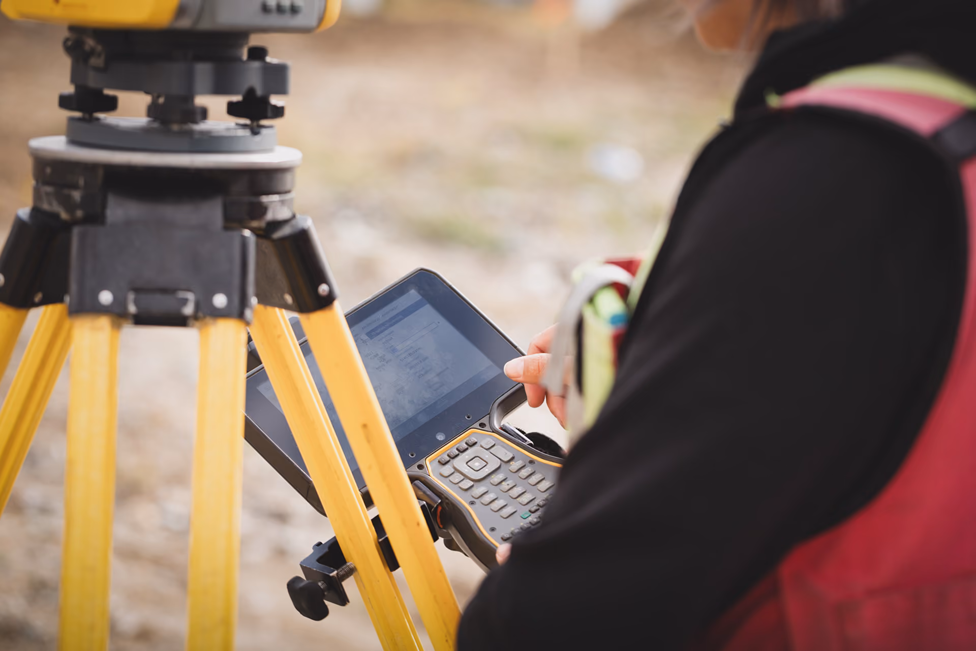 Un technicien en gilet de sécurité rouge utilise un appareil de mesure électronique fixé sur un trépied jaune sur un chantier extérieur.