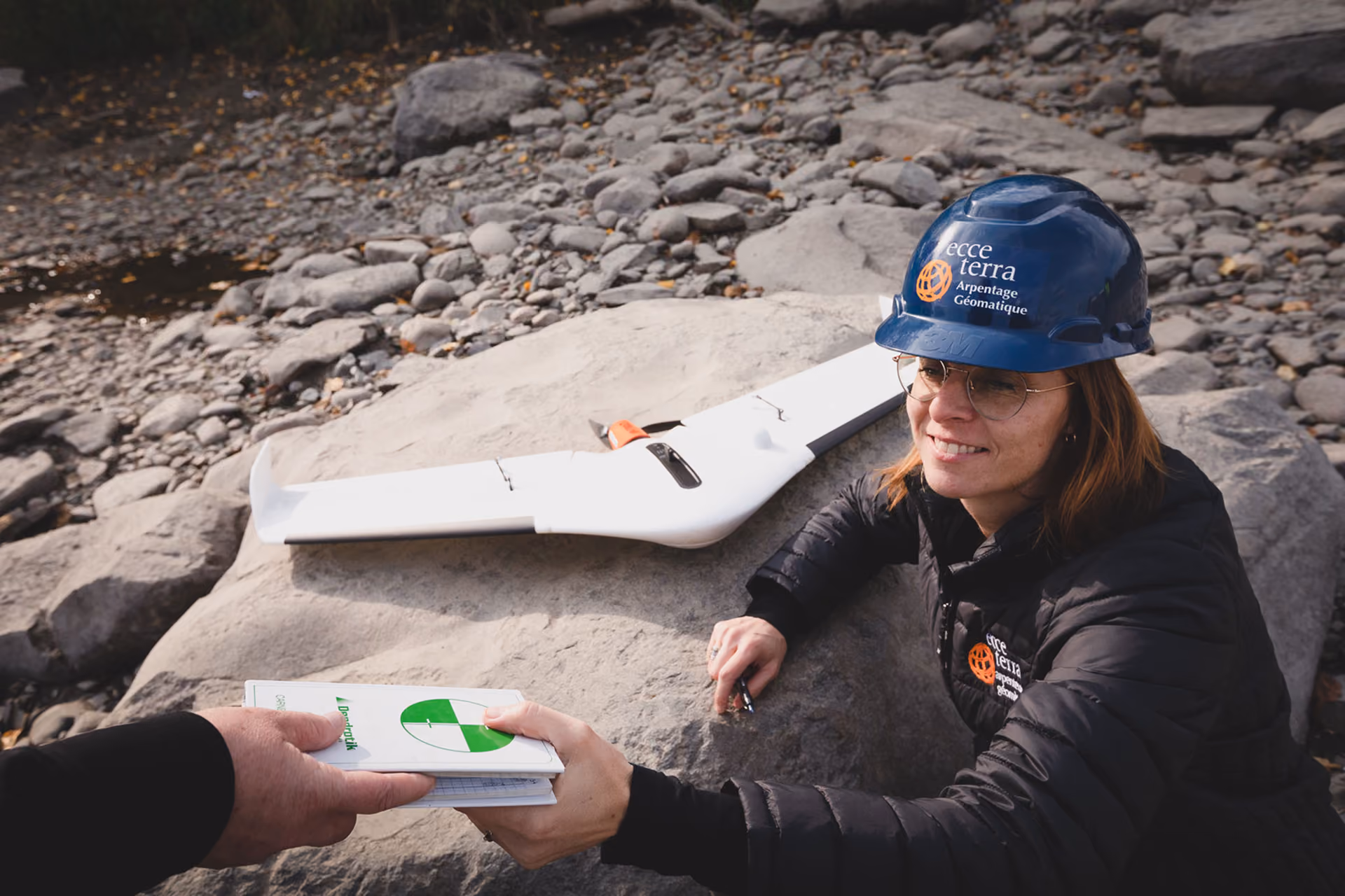 Une femme portant un casque bleu et une veste noire avec le logo 'ecce terra Arpentage Géomatique' sourit et tend un carnet à une autre personne sur un rocher près d'un drone posé sur une grande pierre dans un paysage rocheux.