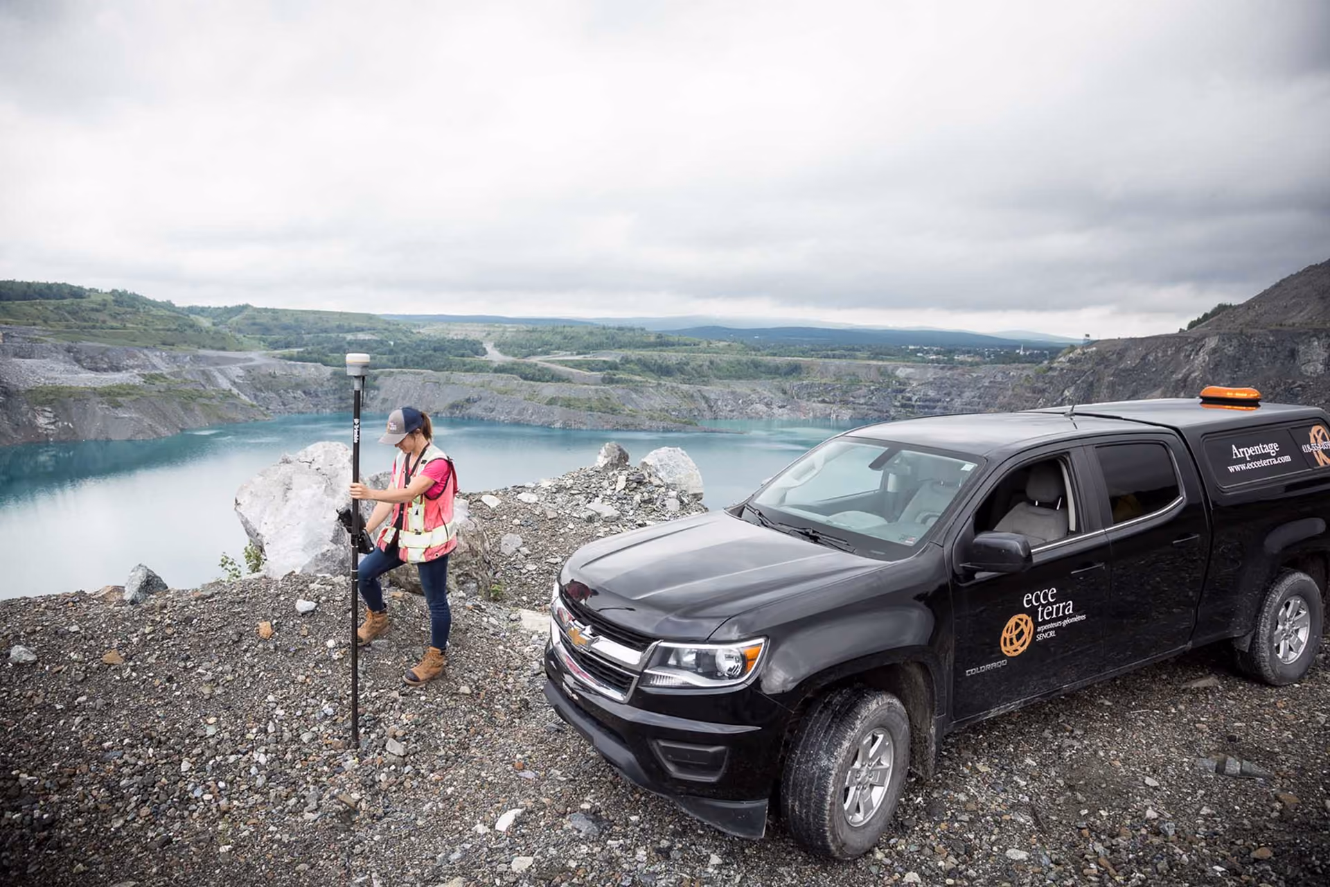 Technicien en arpentage debout sur un terrain rocheux près d'un lac dans une carrière, tenant un équipement de mesure, à côté d'un camion noir marqué Ecce Terra.