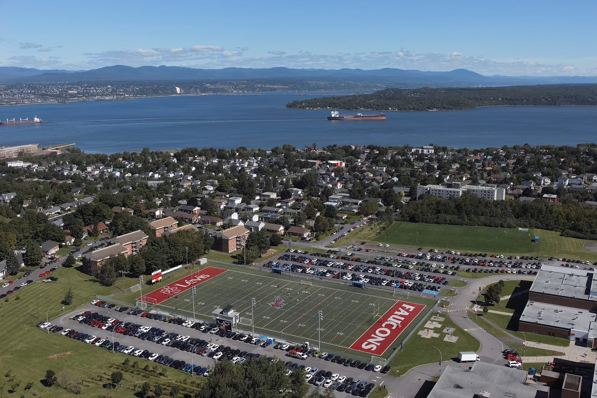 Vue aérienne d'un terrain de football avec les mots 'FALCONS' et 'LEVIS' dans les zones d'en-but, entouré de places de stationnement pleines, avec une ville résidentielle, un grand cours d'eau et des collines en arrière-plan.