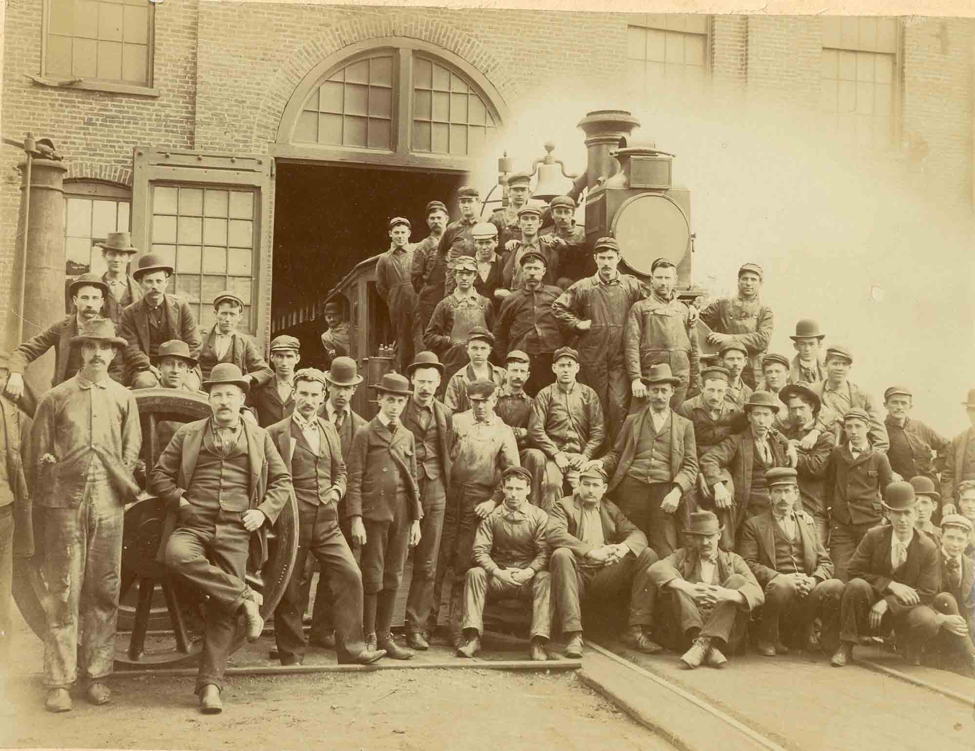 Group portrait of around 45 men posing in front of a train engine and rail tracks at a brick industrial building, with some men standing, some sitting, and others leaning on the train or each other, dressed in early 20th century work clothes and suits with hats.
