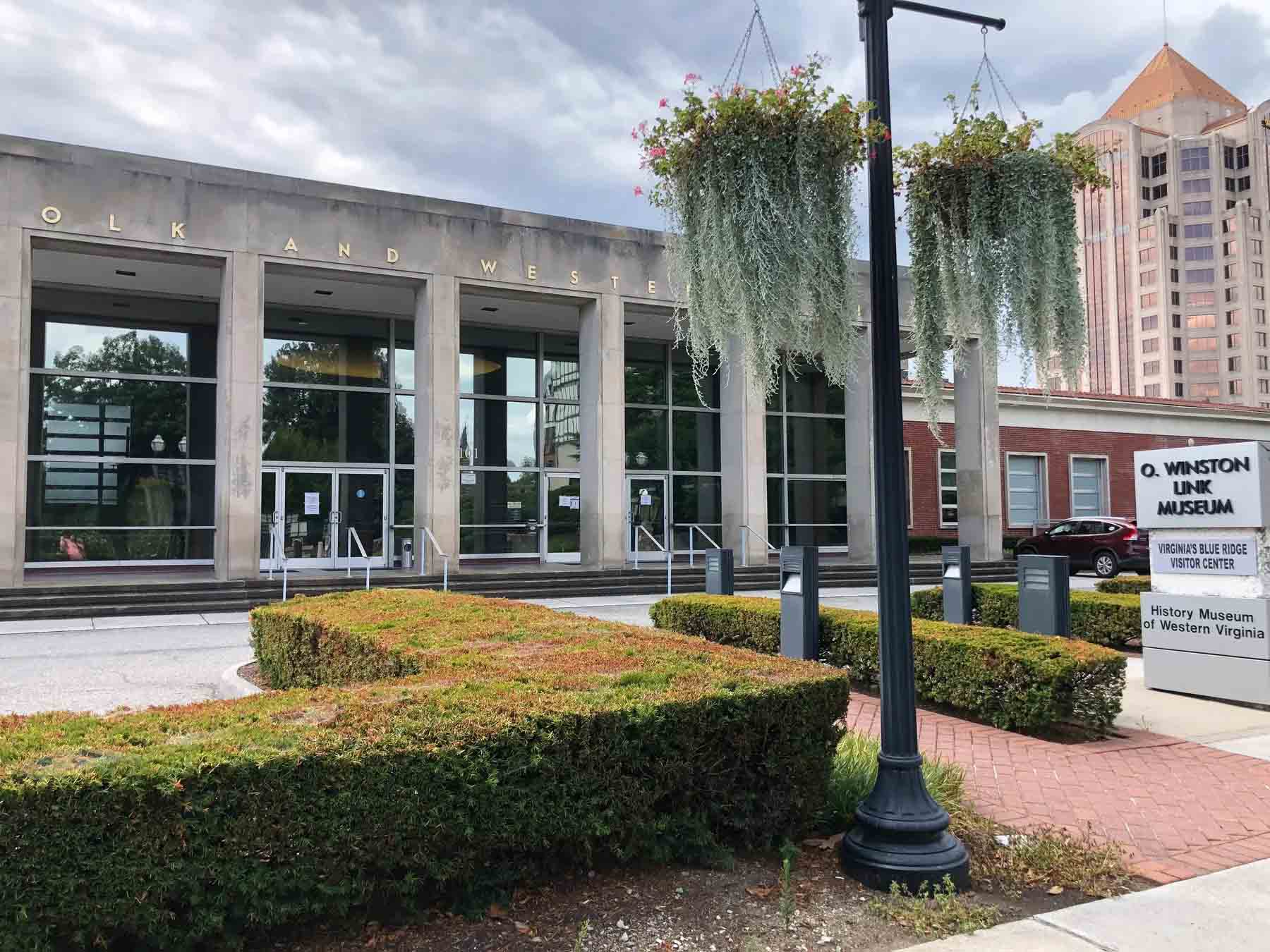 Front entrance of the O. Winston Link Museum and Virginia's Blue Ridge Visitor Center with large glass windows, trimmed bushes, hanging plants, and a tall building in the background.