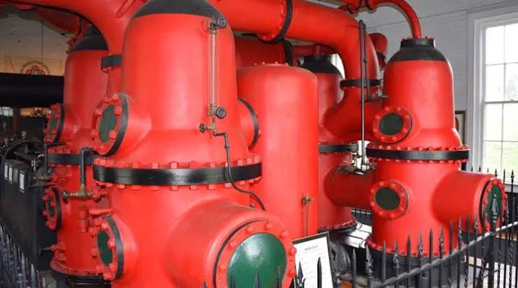 Large red industrial water pump structures with pipes and gauges inside a white room with windows, surrounded by a black metal fence.