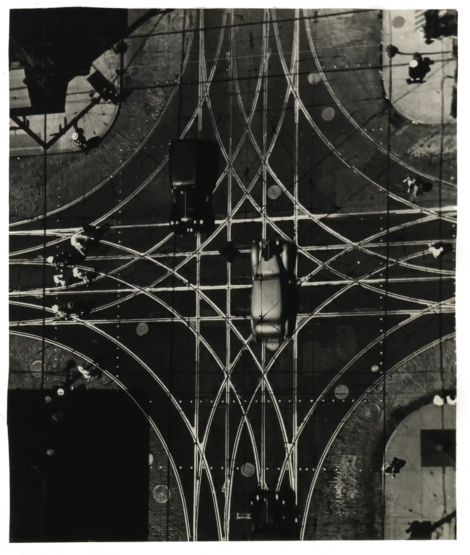 Overhead black and white photo of a traffic intersection featuring multiple tram or trolley tracks crisscrossing, with several vintage cars and pedestrians on sidewalks.