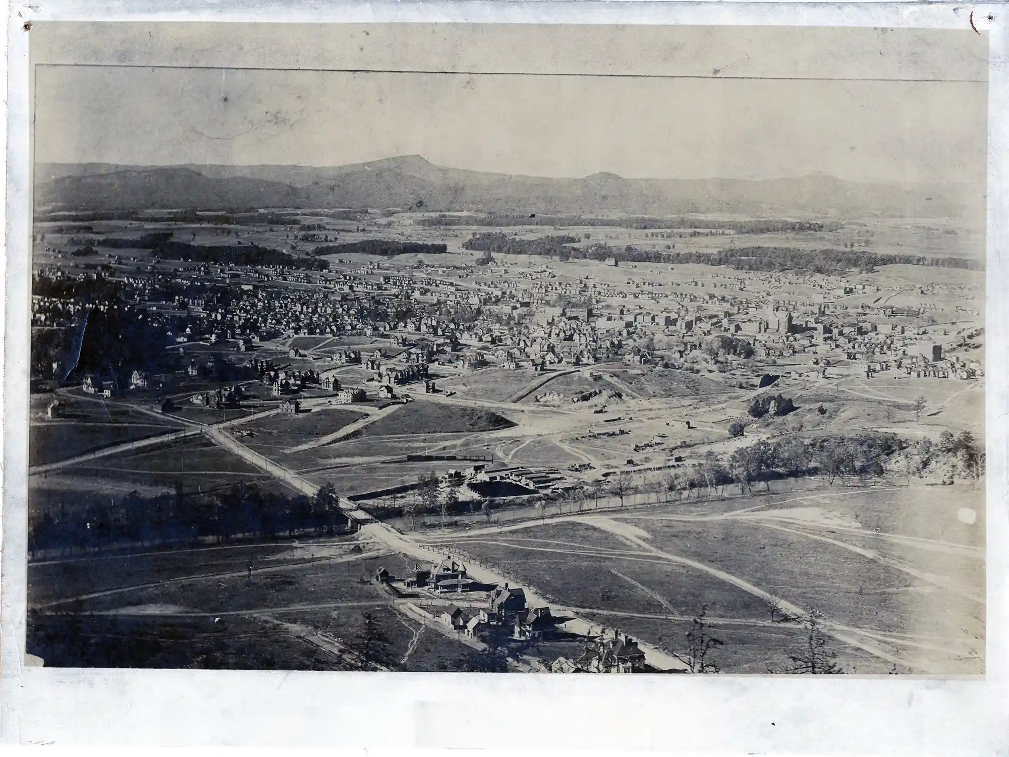 Black and white aerial view of a small town with scattered houses, roads, open fields, and wooded hills in the distance under a clear sky.