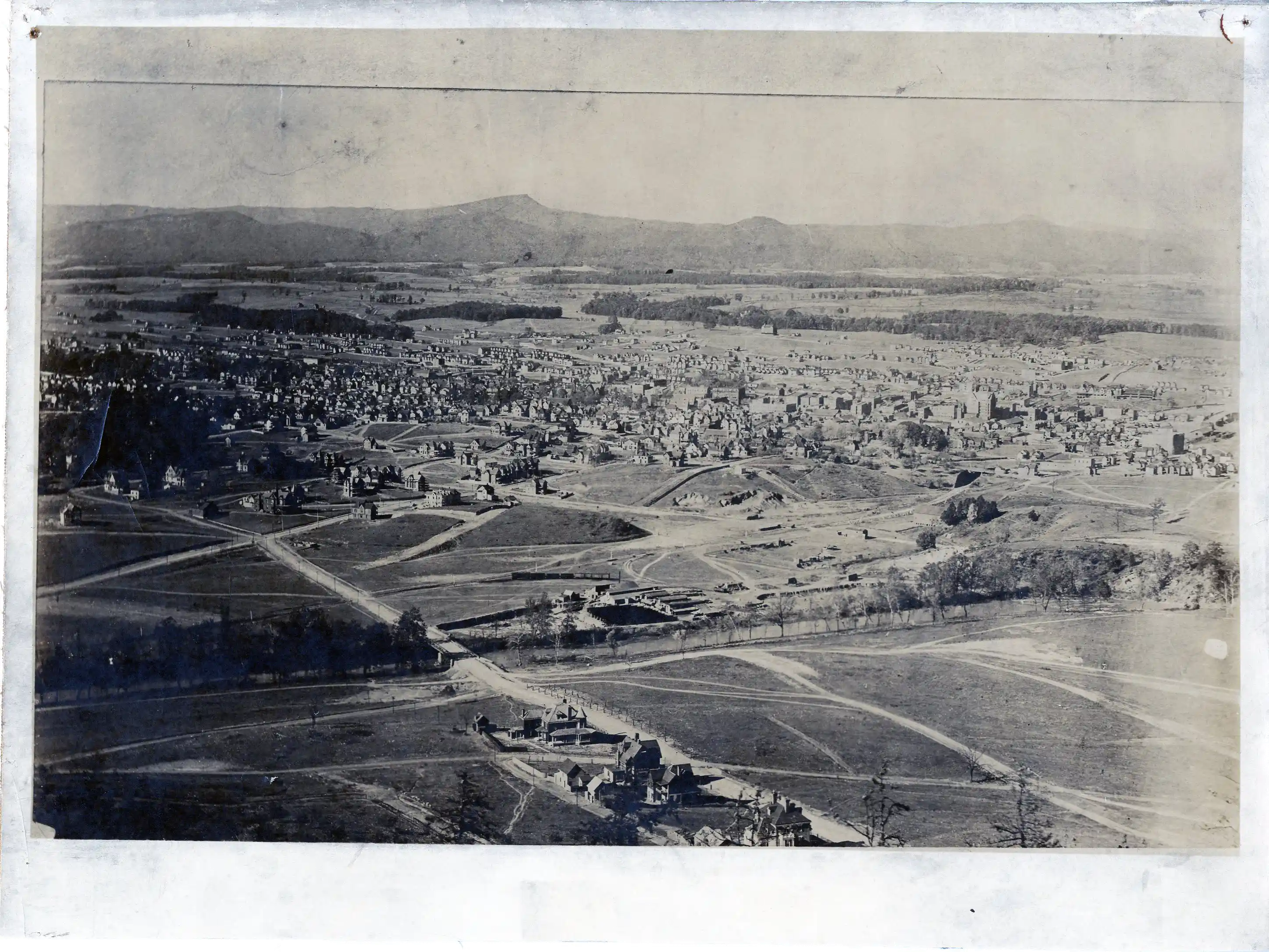 Black-and-white aerial photograph showing an early 20th-century town with numerous houses, roads, open fields, and a mountainous backdrop.