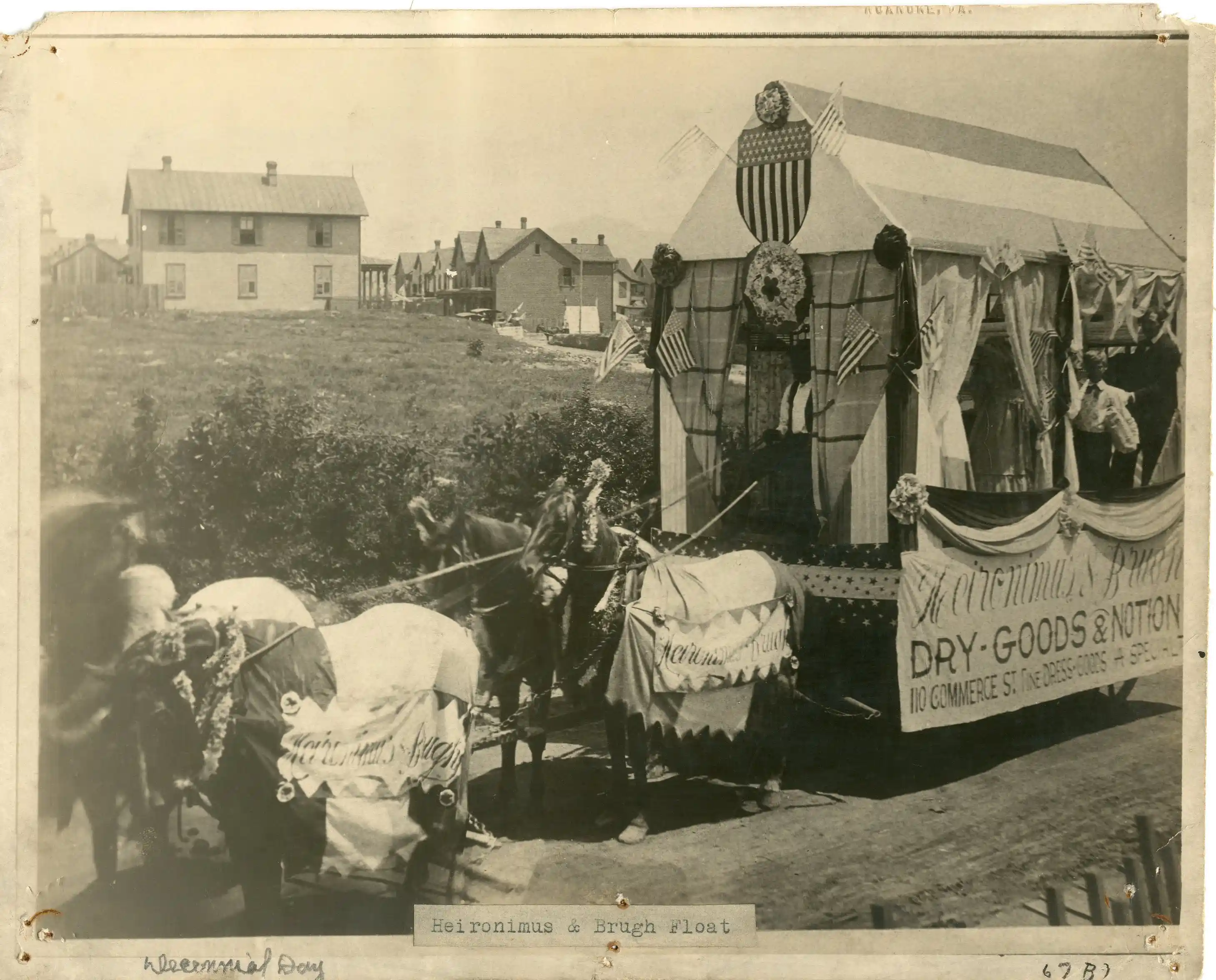 Black and white photo of a horse-drawn parade float decorated with American flags and banners reading 'Heironimus & Brugh DRY GOODS & NOTION 110 Commerce St.' with houses in the background.