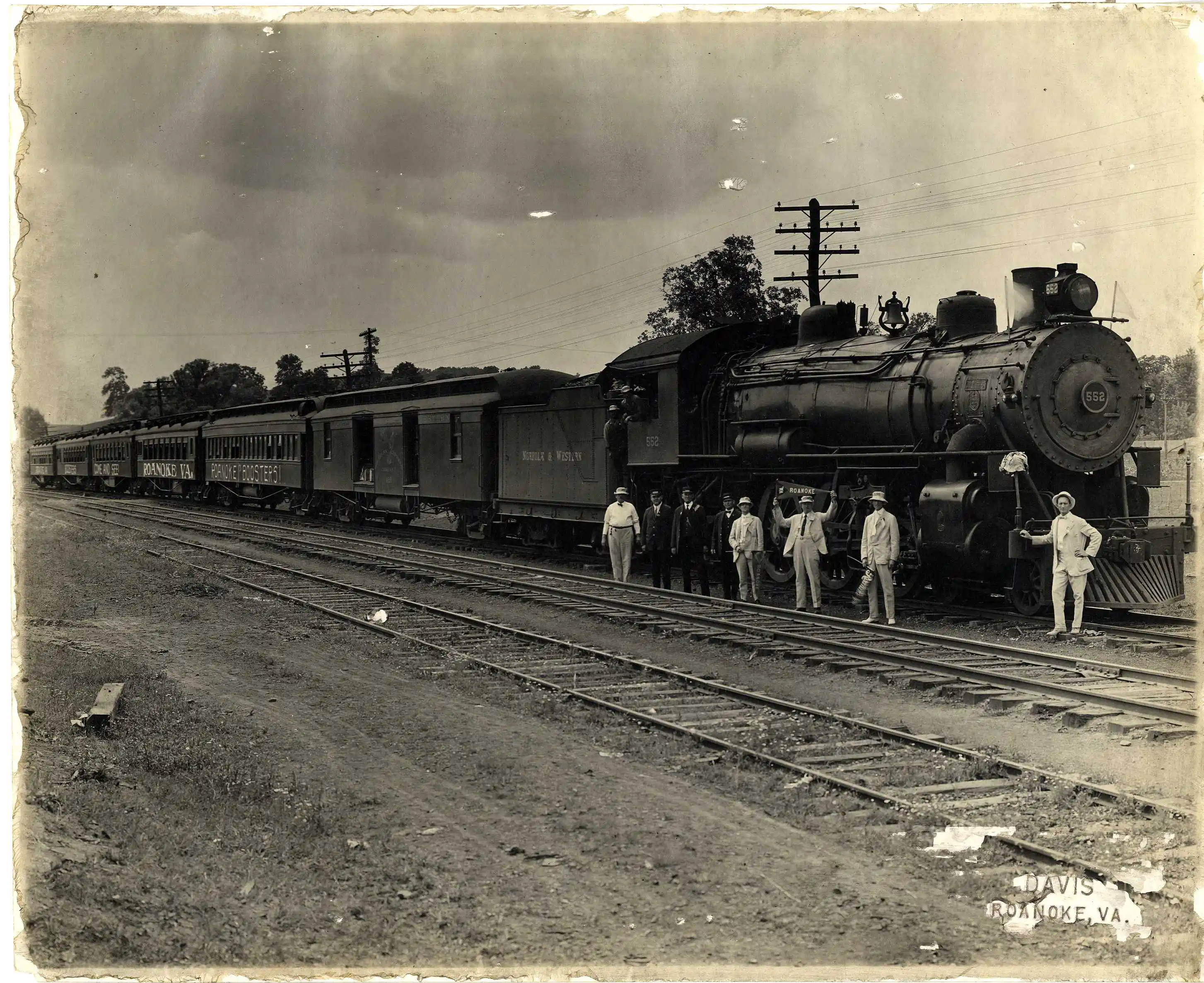 Black and white vintage photograph of a large steam locomotive train with multiple carriages standing on railroad tracks. Nine men dressed in early 20th century attire, including hats and suits, pose along the side of the train. The front of the engine bears the number 552. Trees and telegraph poles are visible in the background. The photograph has an aged, worn border and a label reading 'Davis Roanoke, Va.'