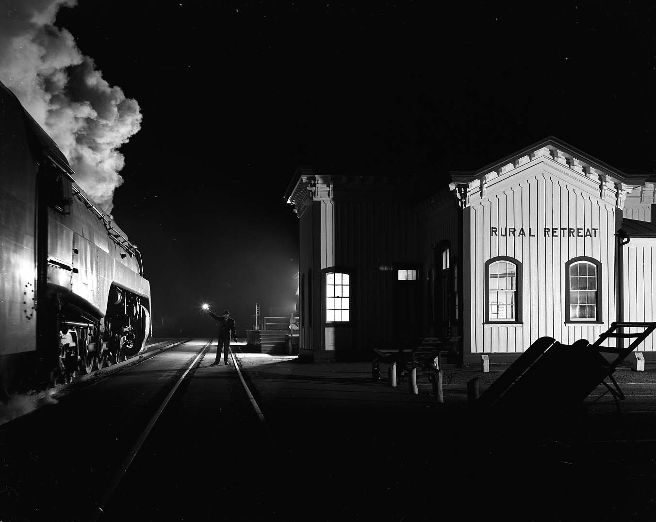 Black and white nighttime scene at Rural Retreat train station with a steam locomotive on the left emitting smoke, and a railroad worker holding a light standing on the tracks near the station building.