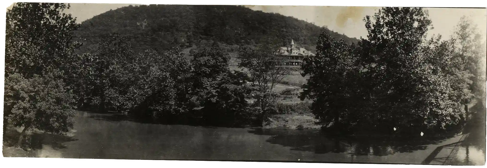 Black and white panoramic photo of a large multi-story building with a steeple-like tower on a hillside surrounded by dense trees, overlooking a calm body of water in the foreground.