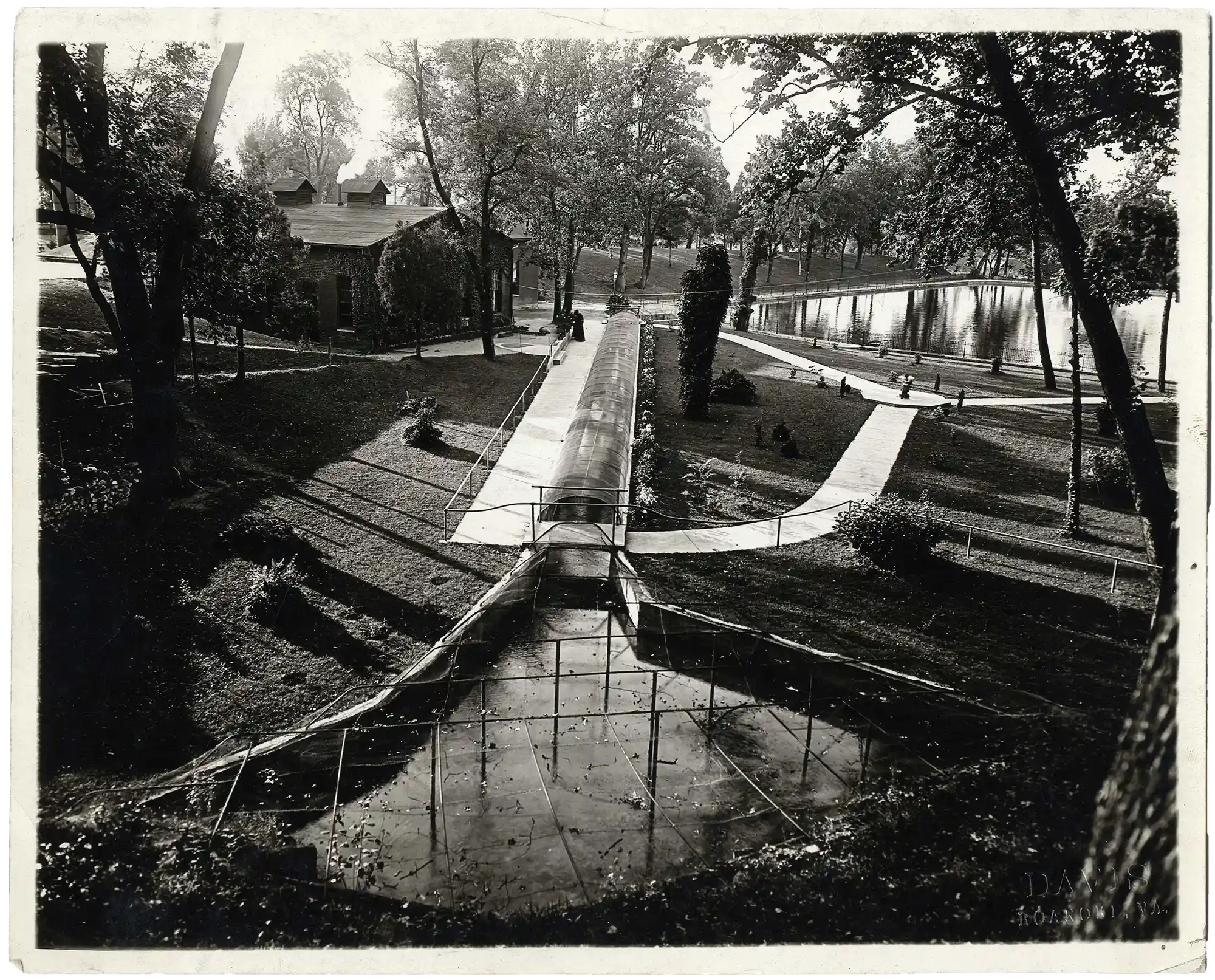 Black and white photo showing a waterway system in a park-like setting with trees, paved paths, and a building partially covered with ivy, with a reflective pond in the background.