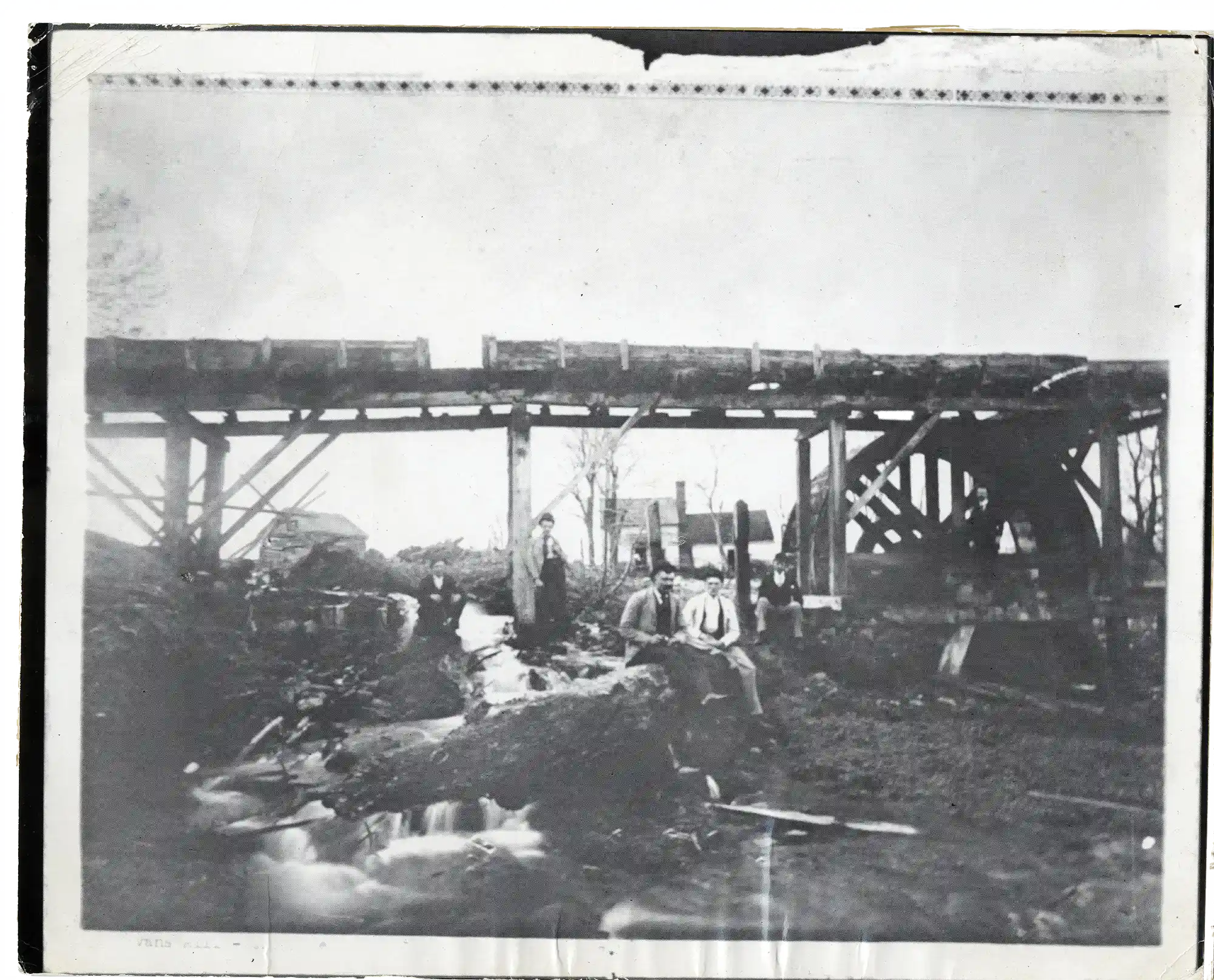 Black and white photo of a wooden water mill with a large wheel beside a small rushing stream, with four men standing or sitting near the structure.