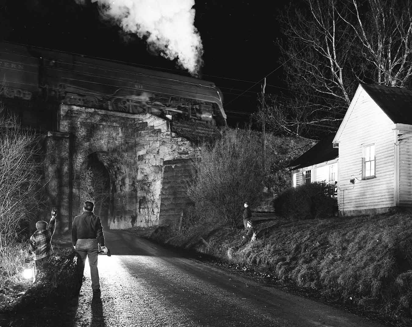 Black and white nighttime scene of four people by a rural road under a small stone railway bridge, watching a steam locomotive pass overhead emitting thick white smoke, with a white house and leafless trees nearby.