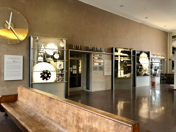 Interior view of the Winston Churchill Museum entrance with large black and white historical photo displays, a wooden bench in the foreground, and a large wall clock to the left on a beige wall.