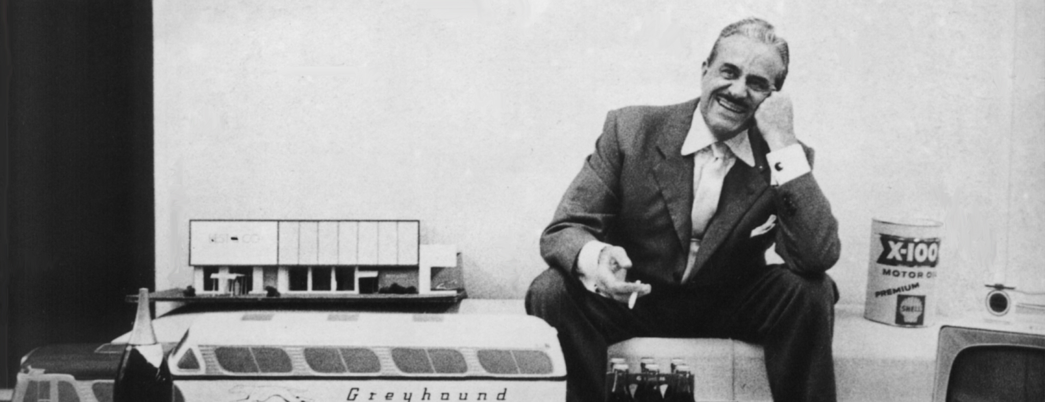 Black and white photo of a smiling man in a suit sitting among miniature models including a Greyhound bus, a building, a bottle, and a can of X-100 motor oil.