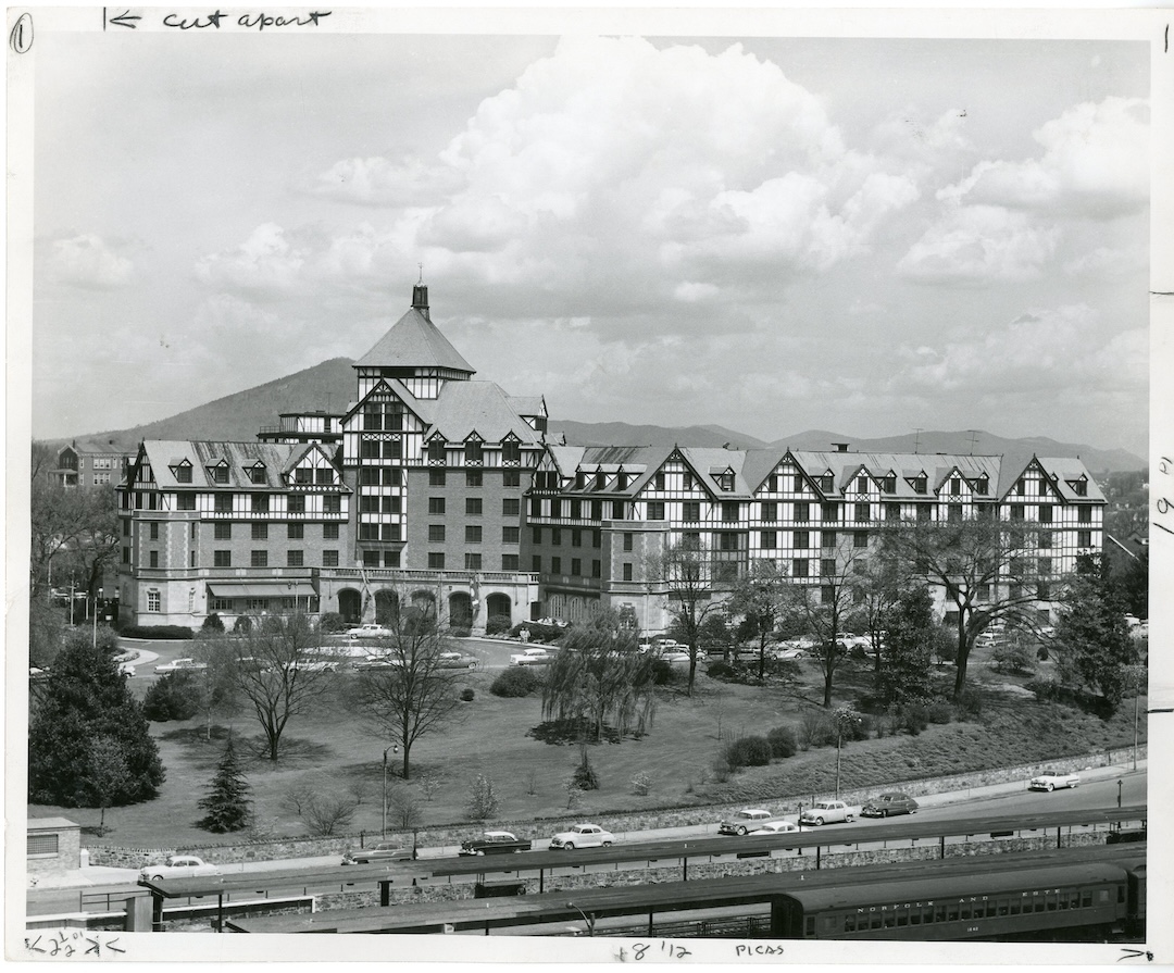 Black and white photograph of a large Tudor-style building with steep gabled roofs and a central tower, surrounded by trees, lawns, parked vintage cars, and a railroad track in the foreground, with hills in the background.