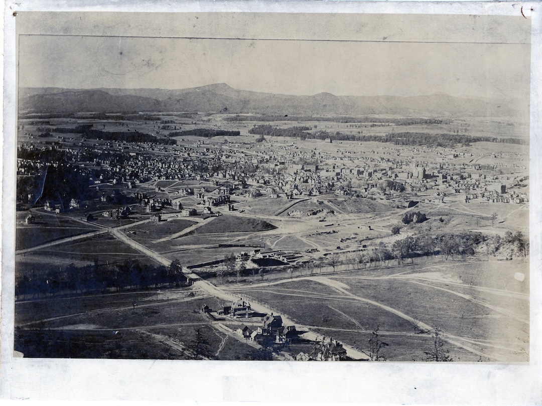 Black and white aerial view of a small town or city from an elevated position, showing a mix of buildings, roads, open fields, and tree lines with hills in the background.