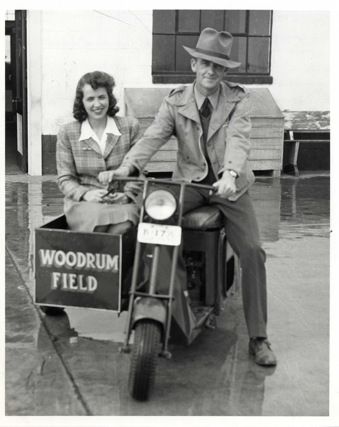 Black and white photo of a man wearing a fedora and trench coat sitting on a small motorcycle with a front sidecar labeled 'WOODRUM FIELD' seating a smiling woman in a plaid jacket and skirt.