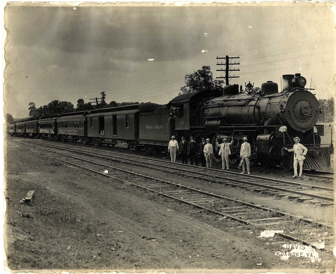 Black and white vintage photo of a steam locomotive train labeled 'Roanoke Booster' on tracks with a group of men standing alongside, some in suits and hats, and one man holding a flag; the setting appears rural with trees and telephone poles in the background. The photo edges are worn and there is a label reading 'DAVIS ROANOKE, VA.'