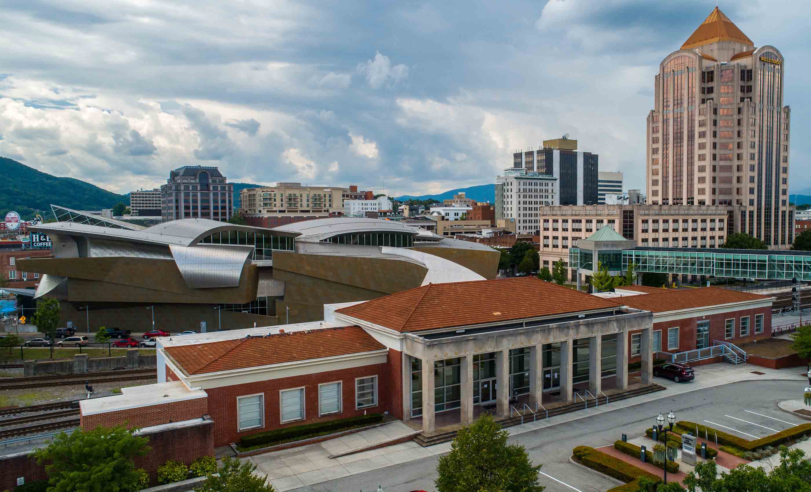 City skyline with a distinctive modern building featuring curved metallic roofs in the midground, a classic red brick building with large windows and columns in the foreground, and a tall office tower labeled Wells Fargo in the background under a cloudy sky.