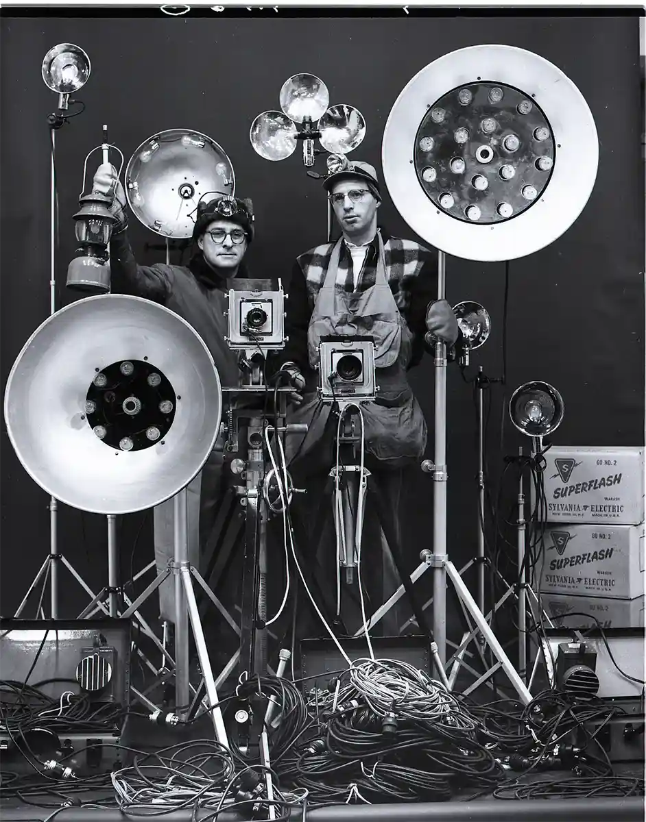 Black and white photo of two men standing among vintage photography equipment, including large studio lights with reflectors, cameras on tripods, and various cables neatly coiled on the floor, with boxes labeled Superflash Sylvania Electric on the right side.