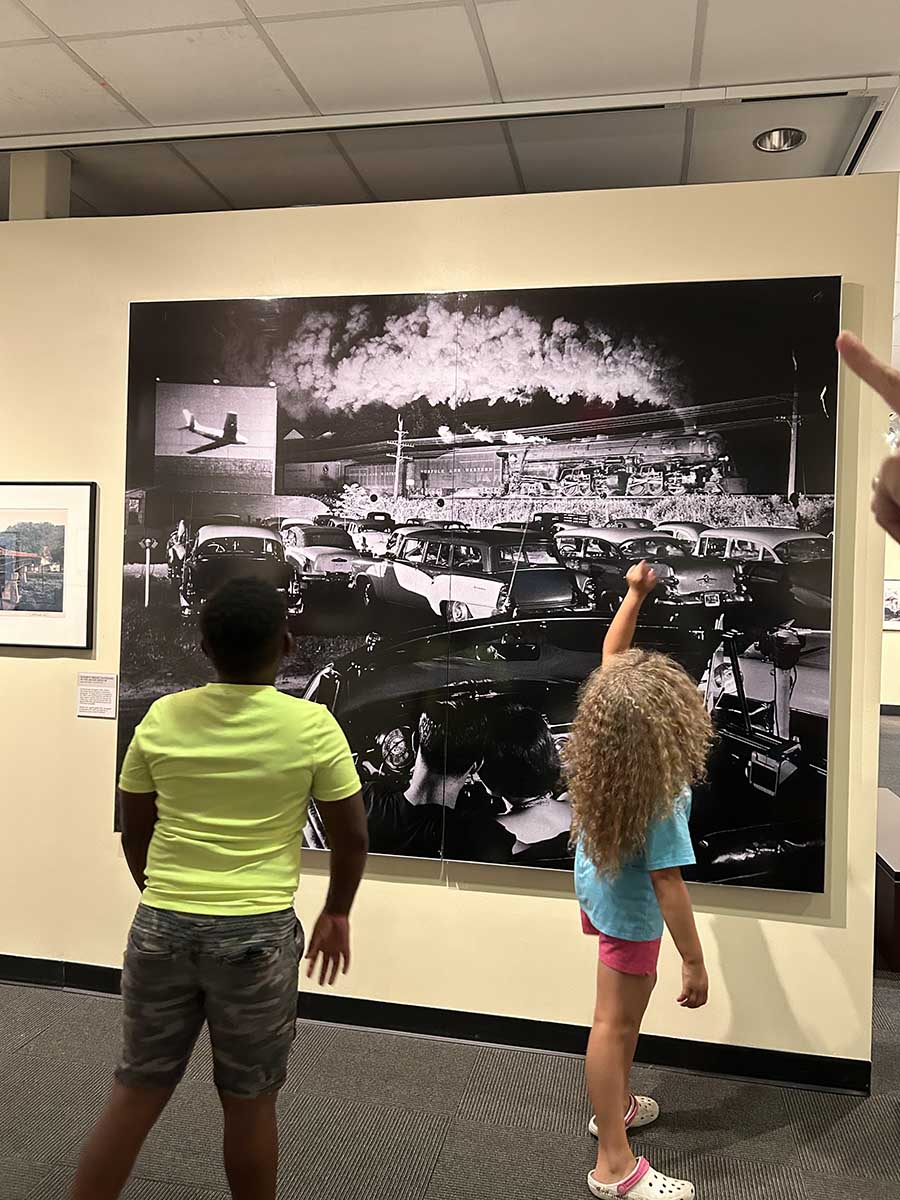 Two children stand in front of a large black-and-white photograph on a museum wall showing a vintage drive-in movie scene with classic cars parked and a steam train passing by in the background under a night sky filled with steam or smoke.