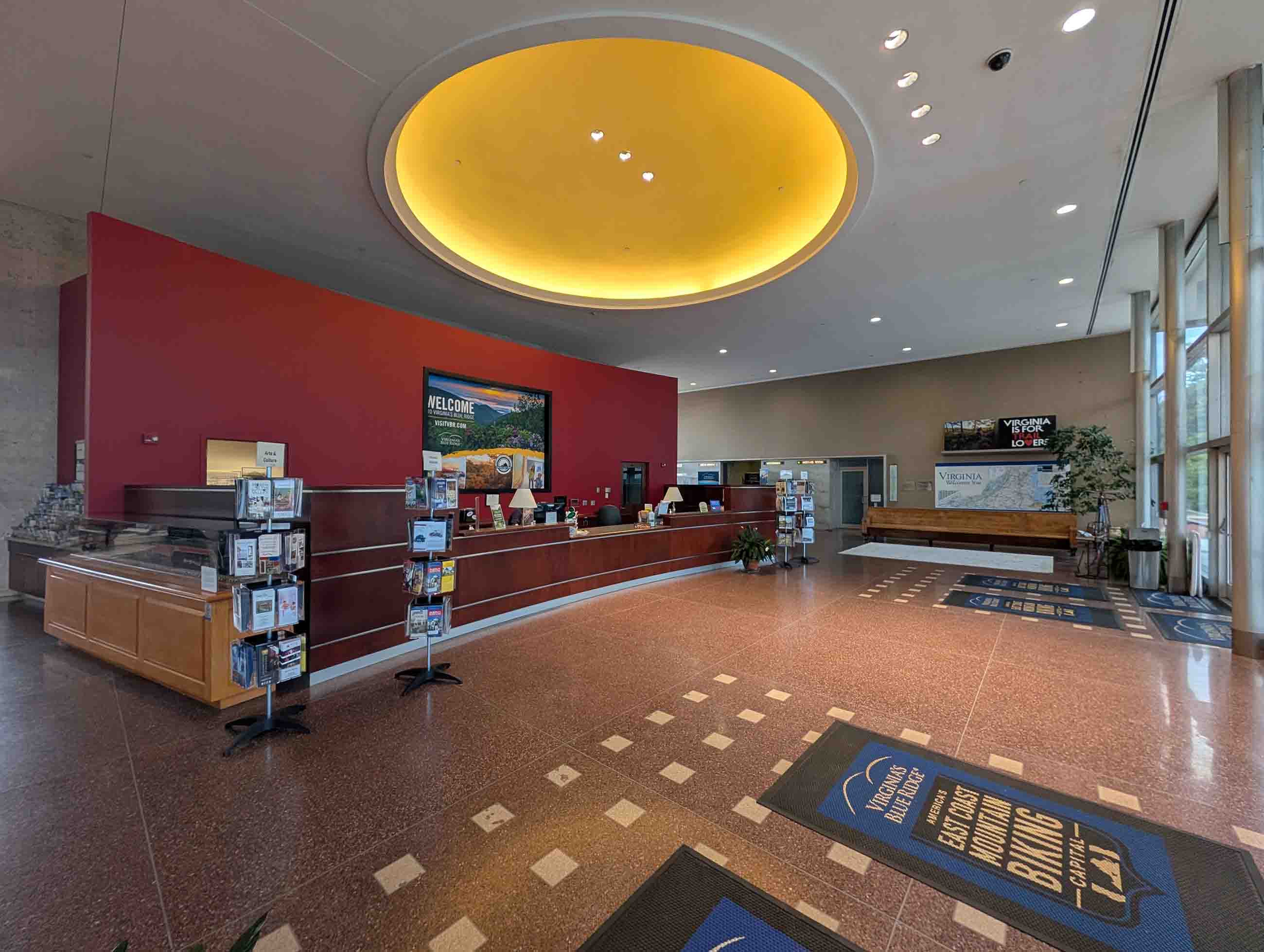 Spacious visitor information center lobby with a curved wooden reception desk against a red wall, brochure stands, a glowing yellow circular ceiling light, large windows, and floor mats reading 'Virginia's Blue Ridge: America's East Coast Mountain Biking Capital.'