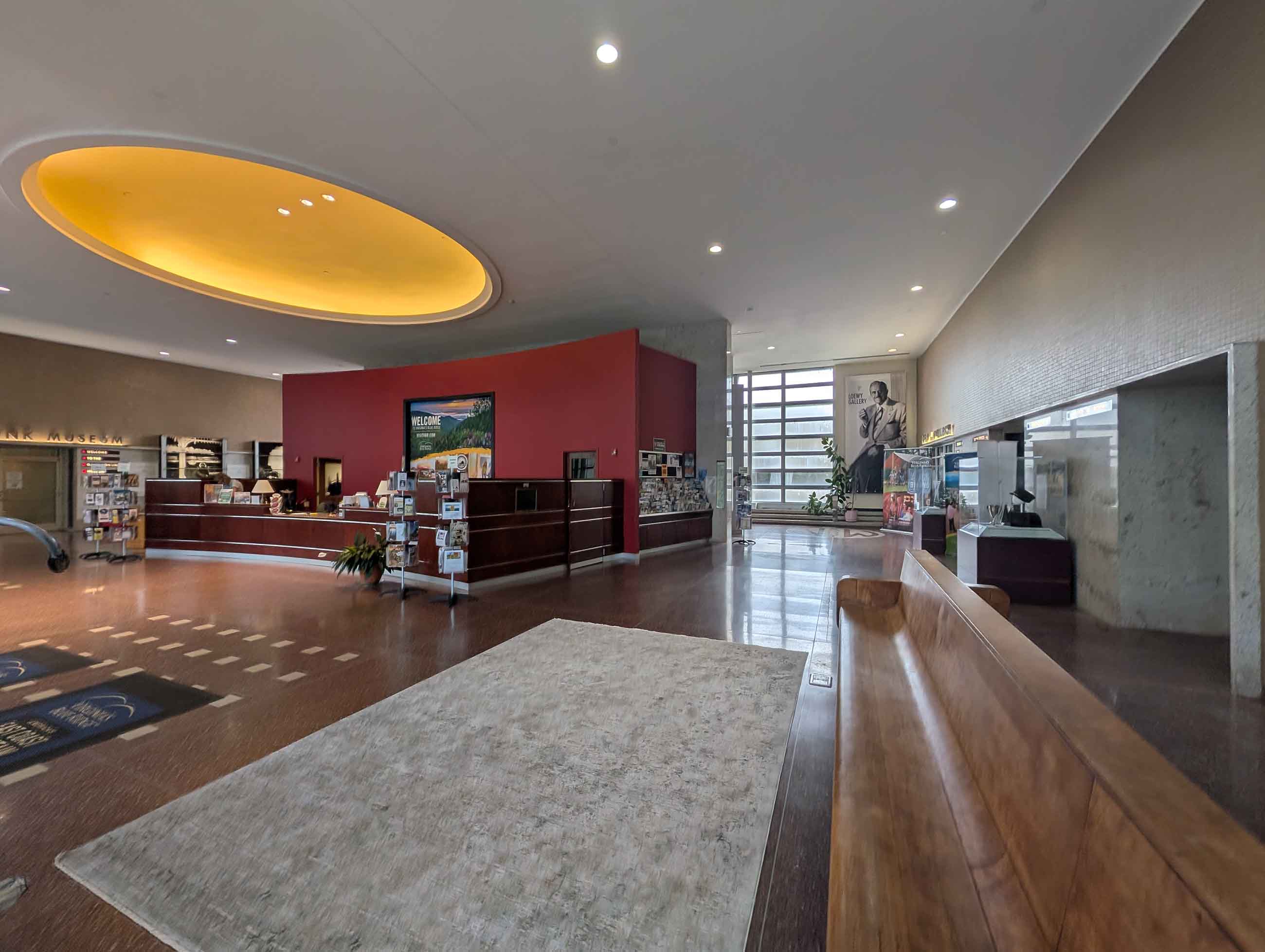 Spacious museum lobby with large circular ceiling light, reception desk against a red wall, seating bench, brochure stands, and a large black and white portrait near the entrance windows.