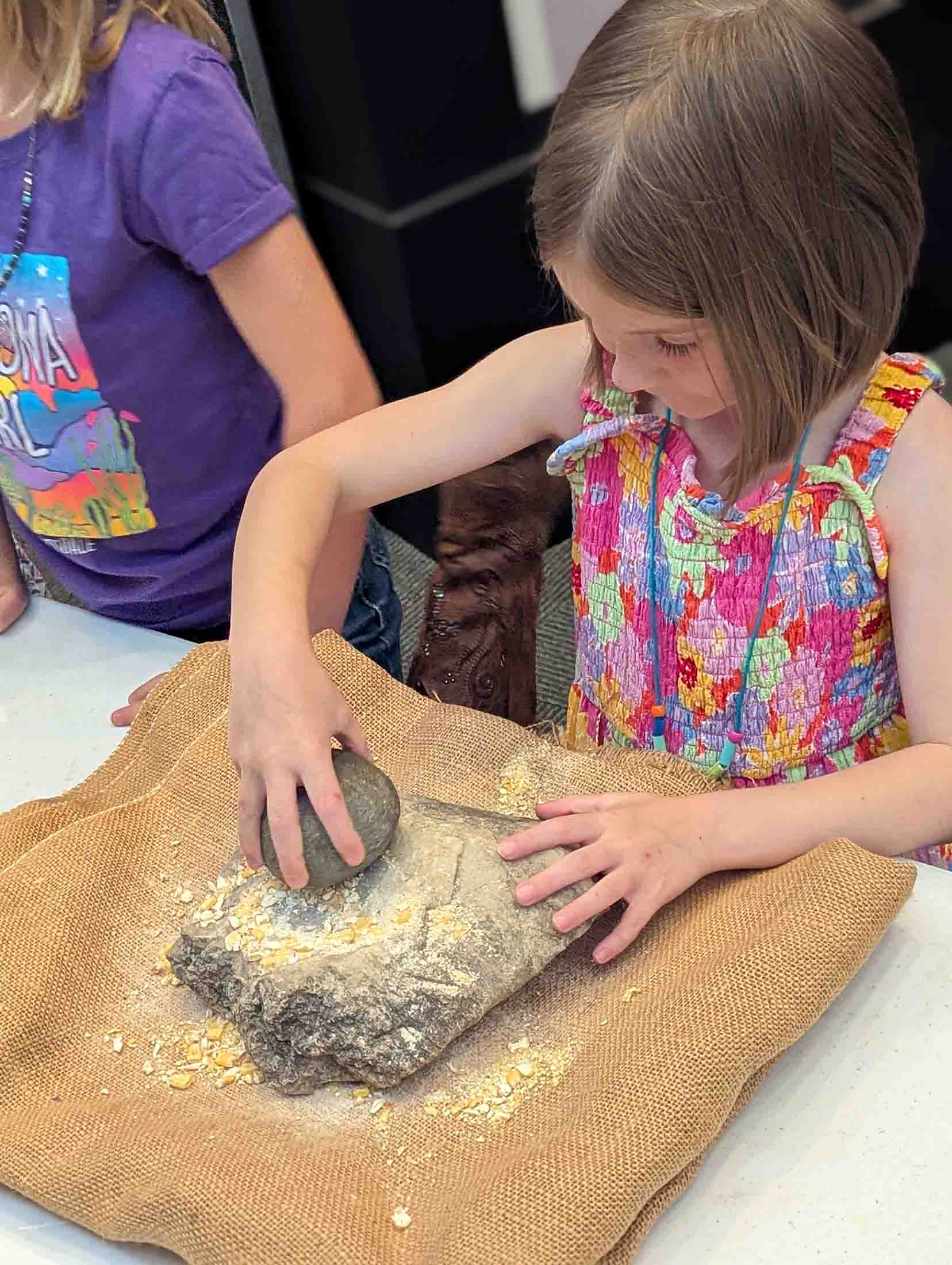 Young girl in a colorful dress grinding dried corn kernels on a large flat stone using a smaller round stone, with corn pieces and powder scattered on a burlap cloth.