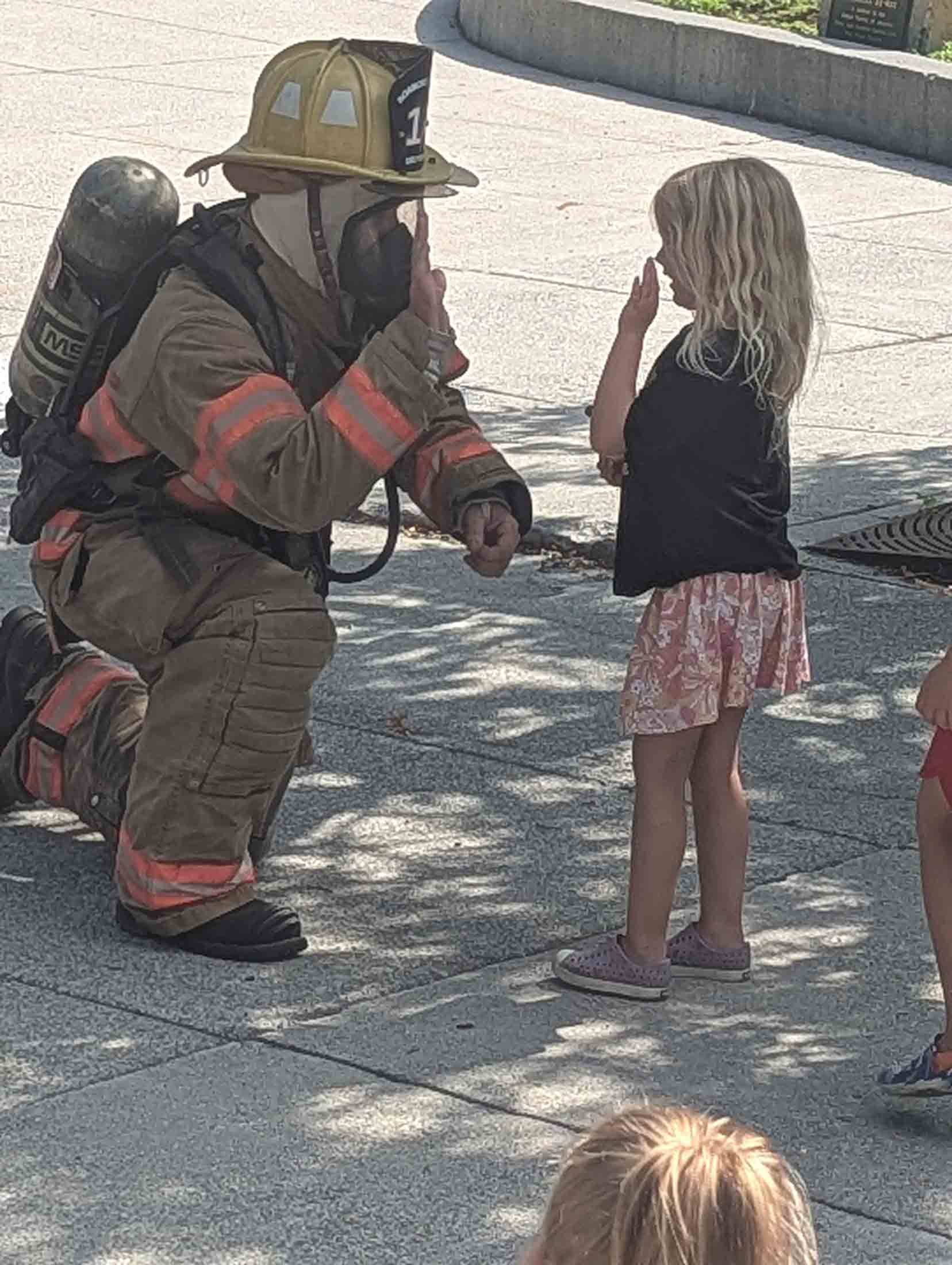 A firefighter in full gear kneels on one knee on a sidewalk, giving a high-five to a young girl with blonde hair who is standing and facing him with her hand raised.