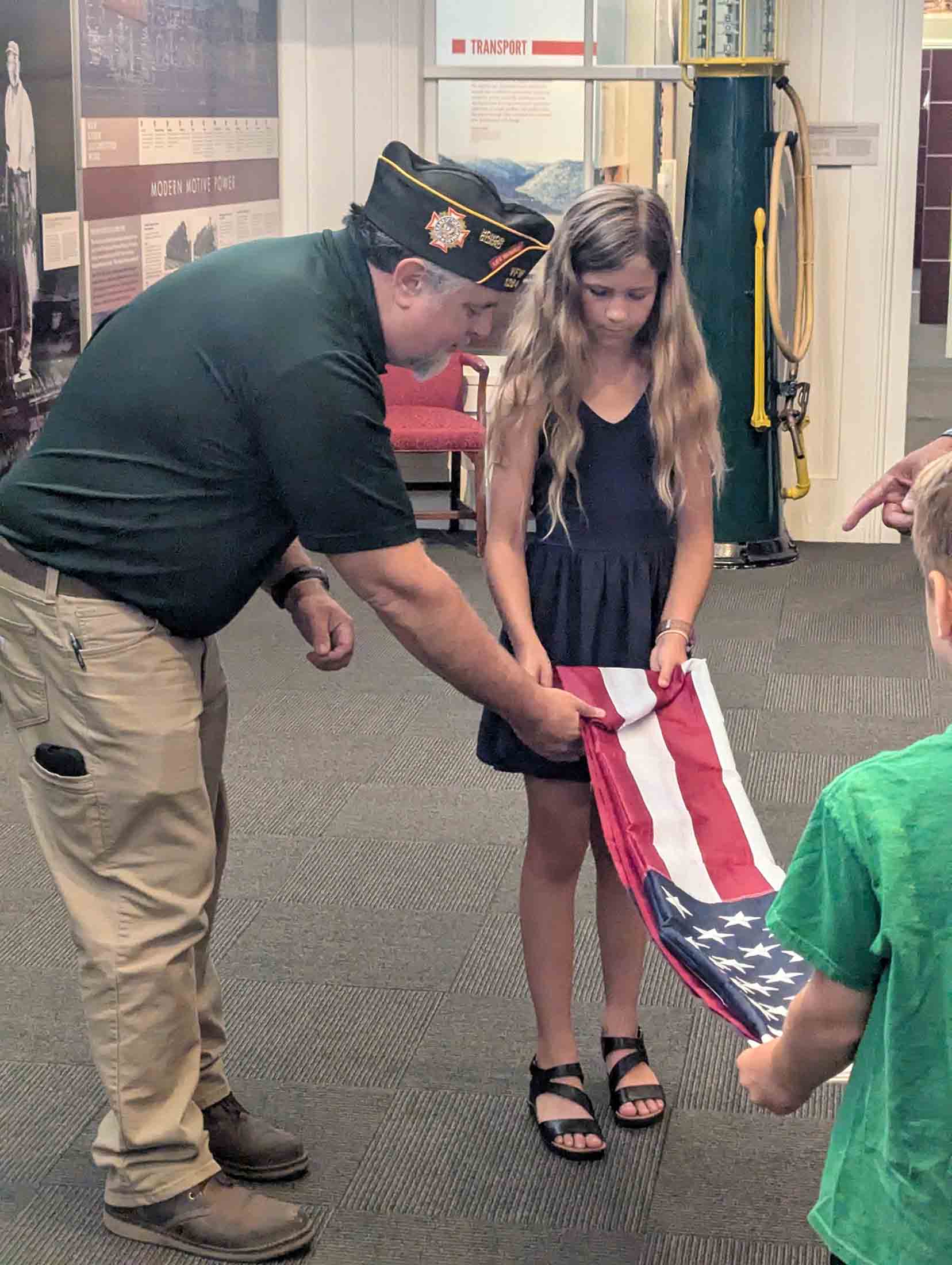Man in a black shirt and VFW cap shows a folded American flag to a young girl in a dark dress while a boy in a green shirt holds the other end of the flag inside a museum or exhibit space.