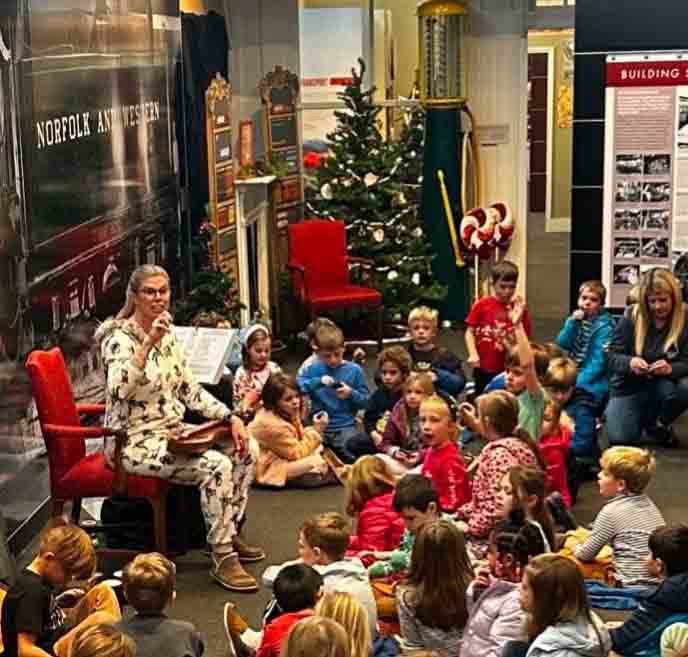 A woman in pajamas sits on a red chair reading a book to a group of children seated on the floor in a cozy indoor setting with Christmas trees and decorations, creating a festive storytime atmosphere.