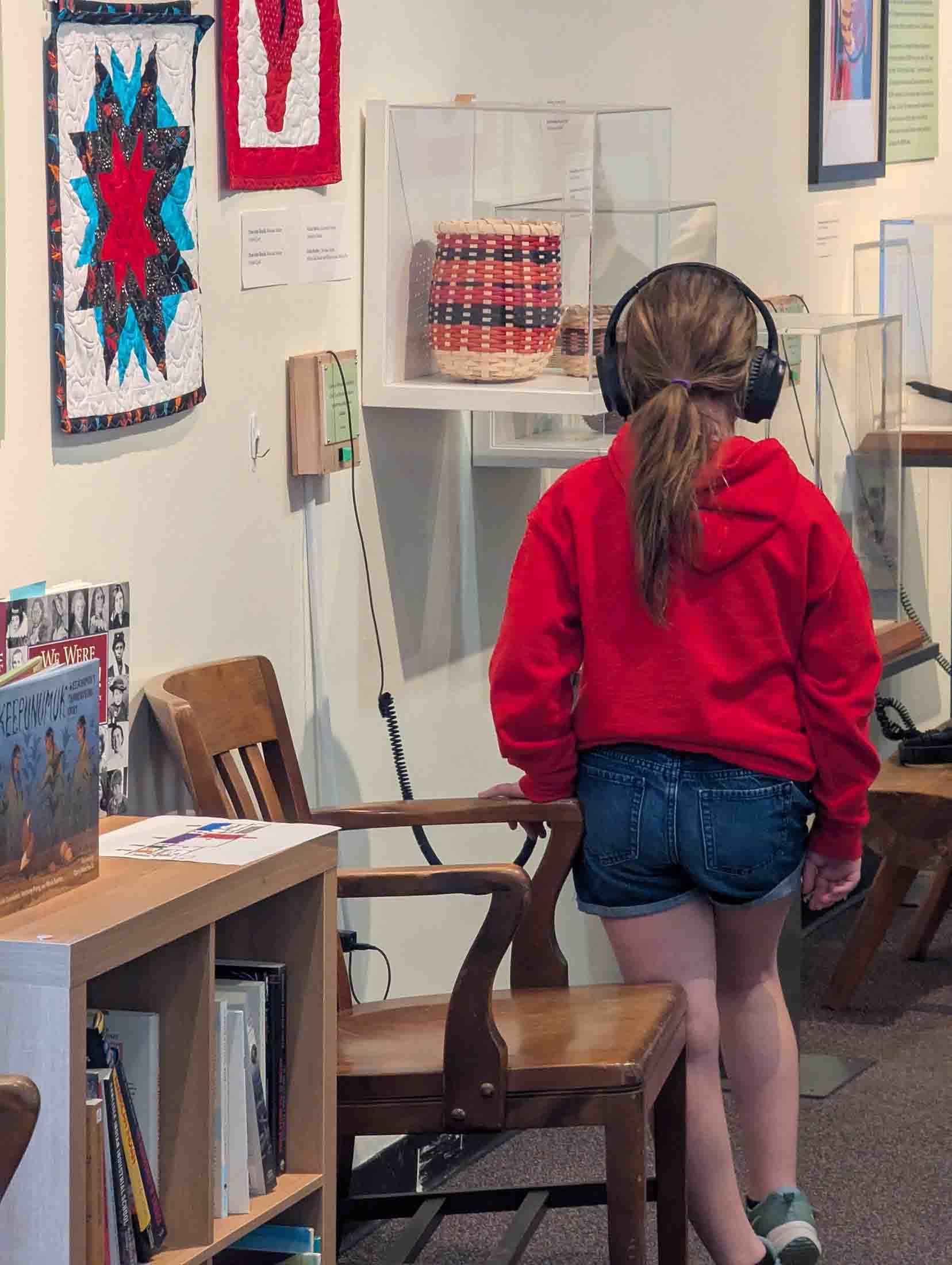 A child wearing a red hoodie, denim shorts, and headphones stands in a museum, facing a wall display case with woven baskets and quilted fabric art pieces.