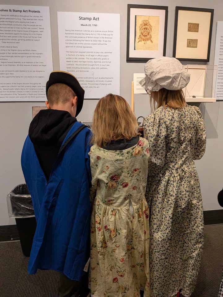 Three children dressed in colonial-era costumes facing a museum wall exhibit about the Stamp Act dated March 22, 1765, with framed documents and informational text panels.