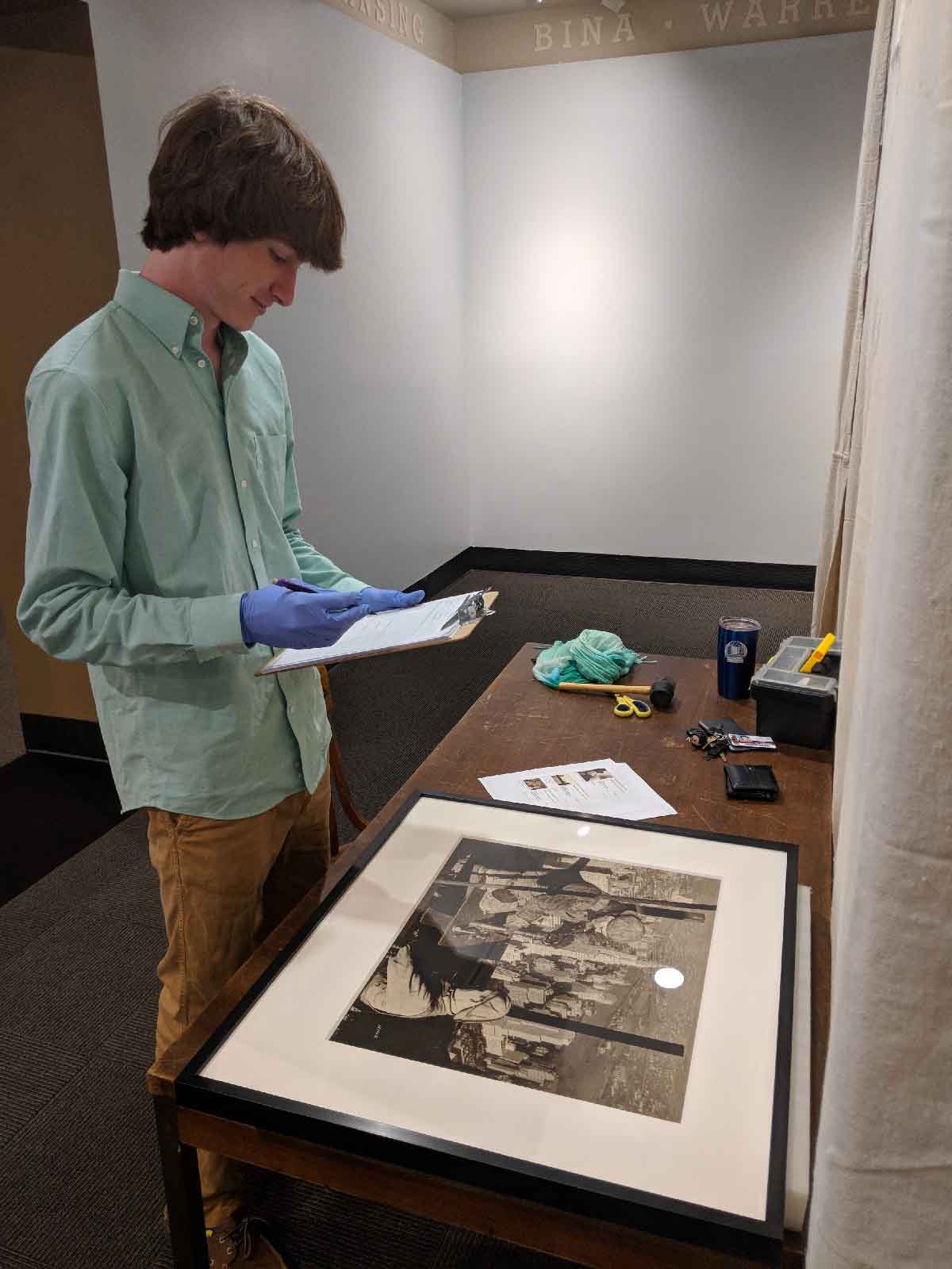 A young man wearing a light green shirt and blue gloves stands by a wooden table, examining and writing on a clipboard. The table holds a framed black-and-white portrait, some papers, a pair of scissors, a black wallet, keys, a tumbler, a black toolbox, and a green cloth. The setting appears to be a museum or gallery space with plain walls.
