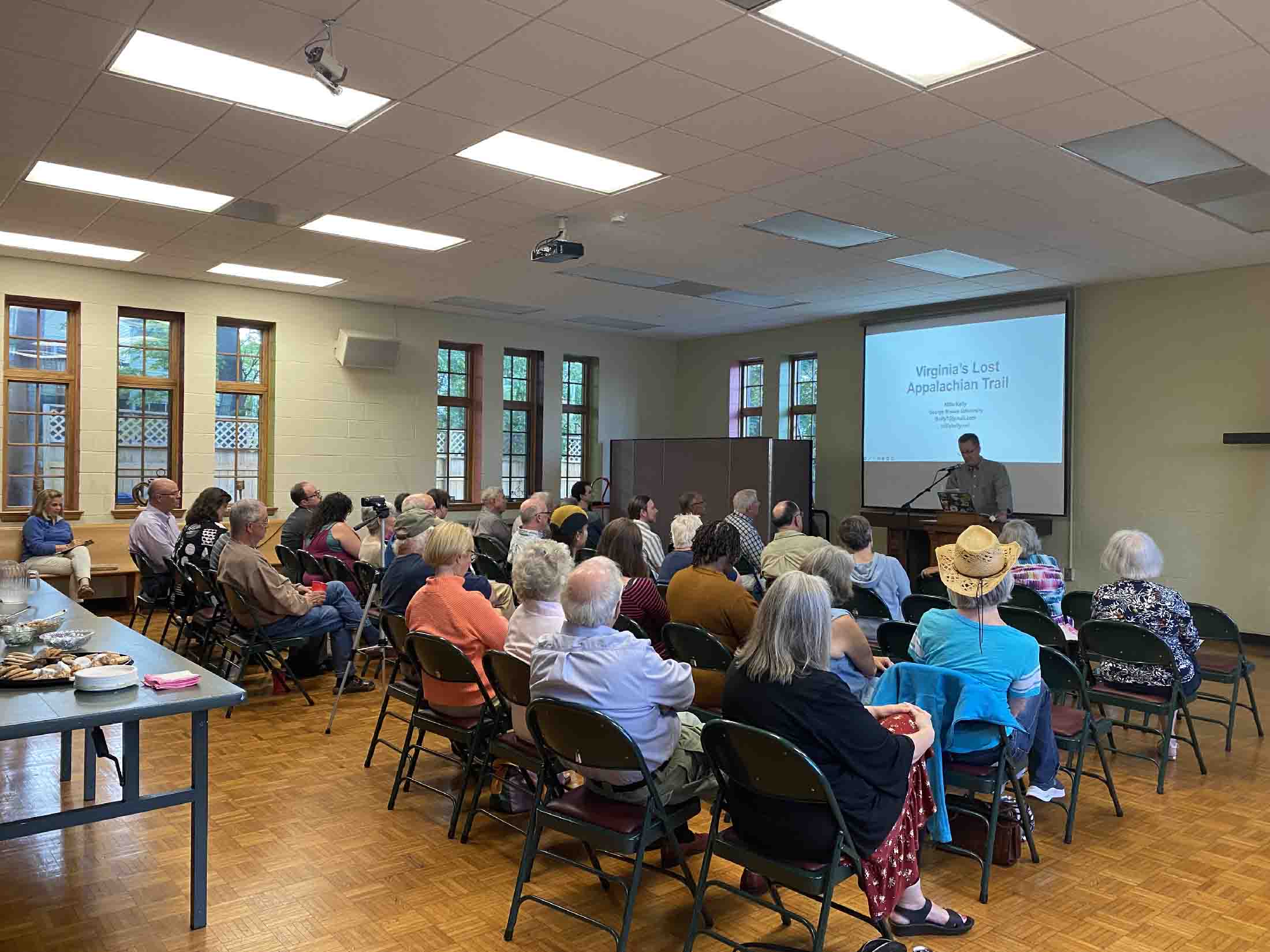 An audience seated in rows in a room listening to a speaker standing at a podium next to a projection screen displaying a presentation titled 'Virginia's Lost Appalachian Trail.'