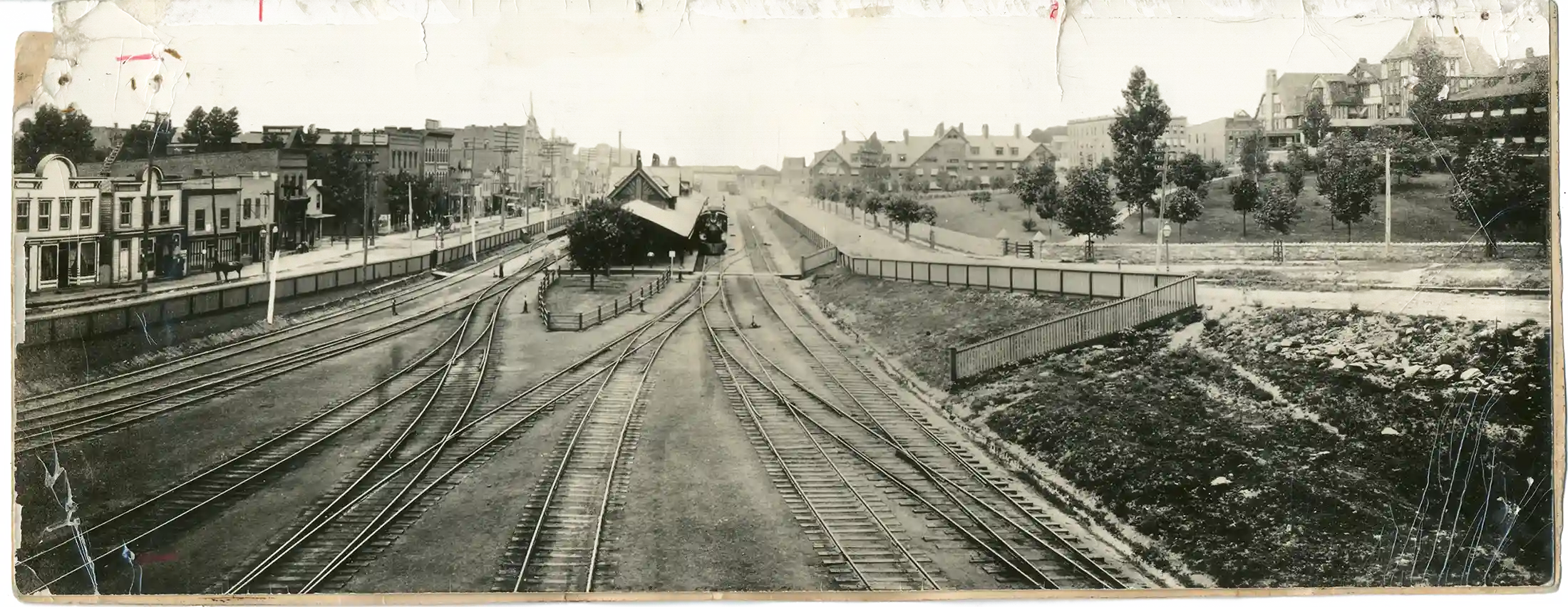 Vintage black and white panoramic photo of an urban railway junction with multiple train tracks diverging, flanked by low-rise buildings on the left and a grassy area with houses on the right, under a cloudy sky.